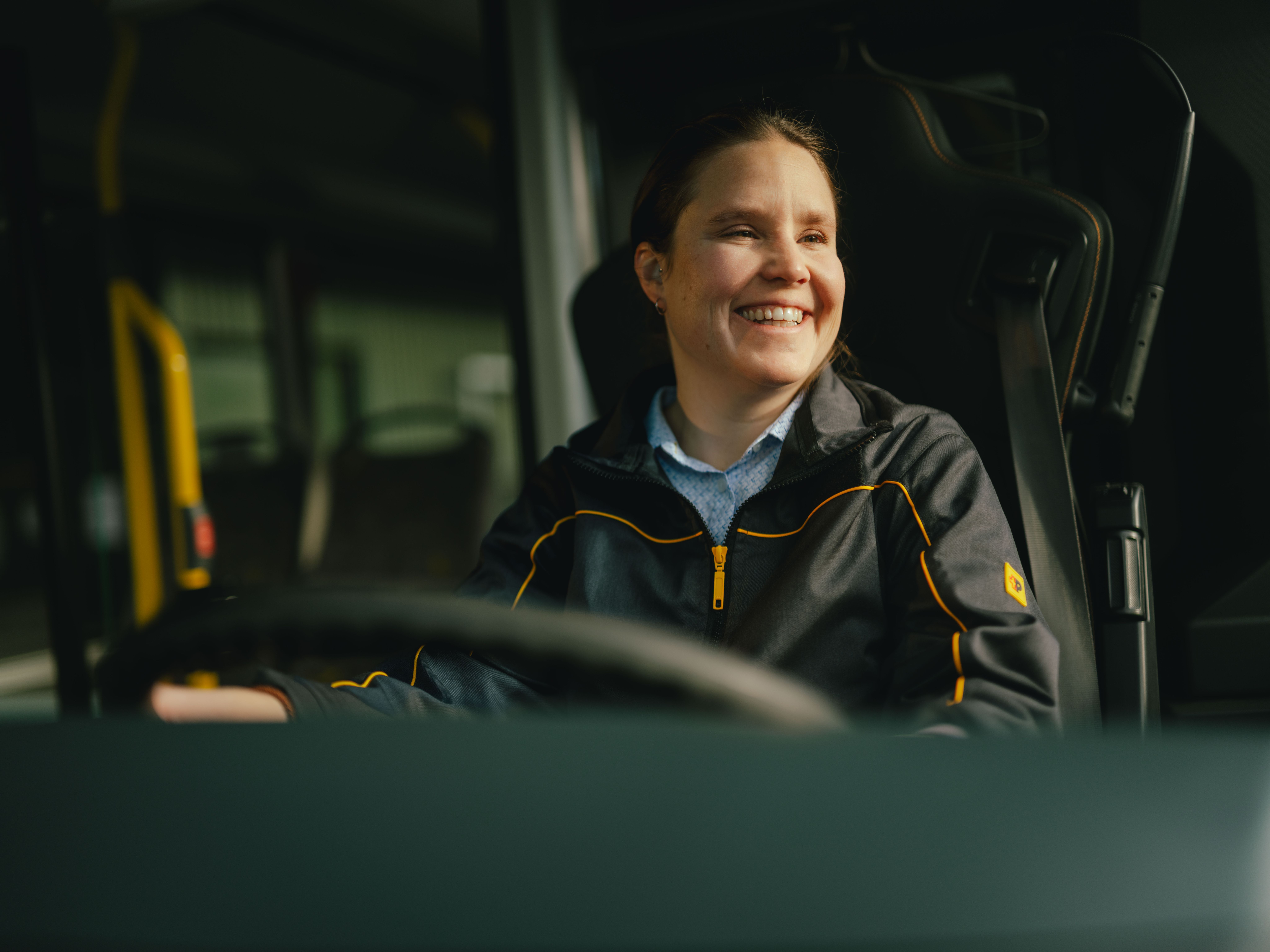 Une personne est assise au volant d’un véhicule, portant une veste foncée avec des accents jaunes. L’intérieur du véhicule, y compris le volant et des poignées, est visible.