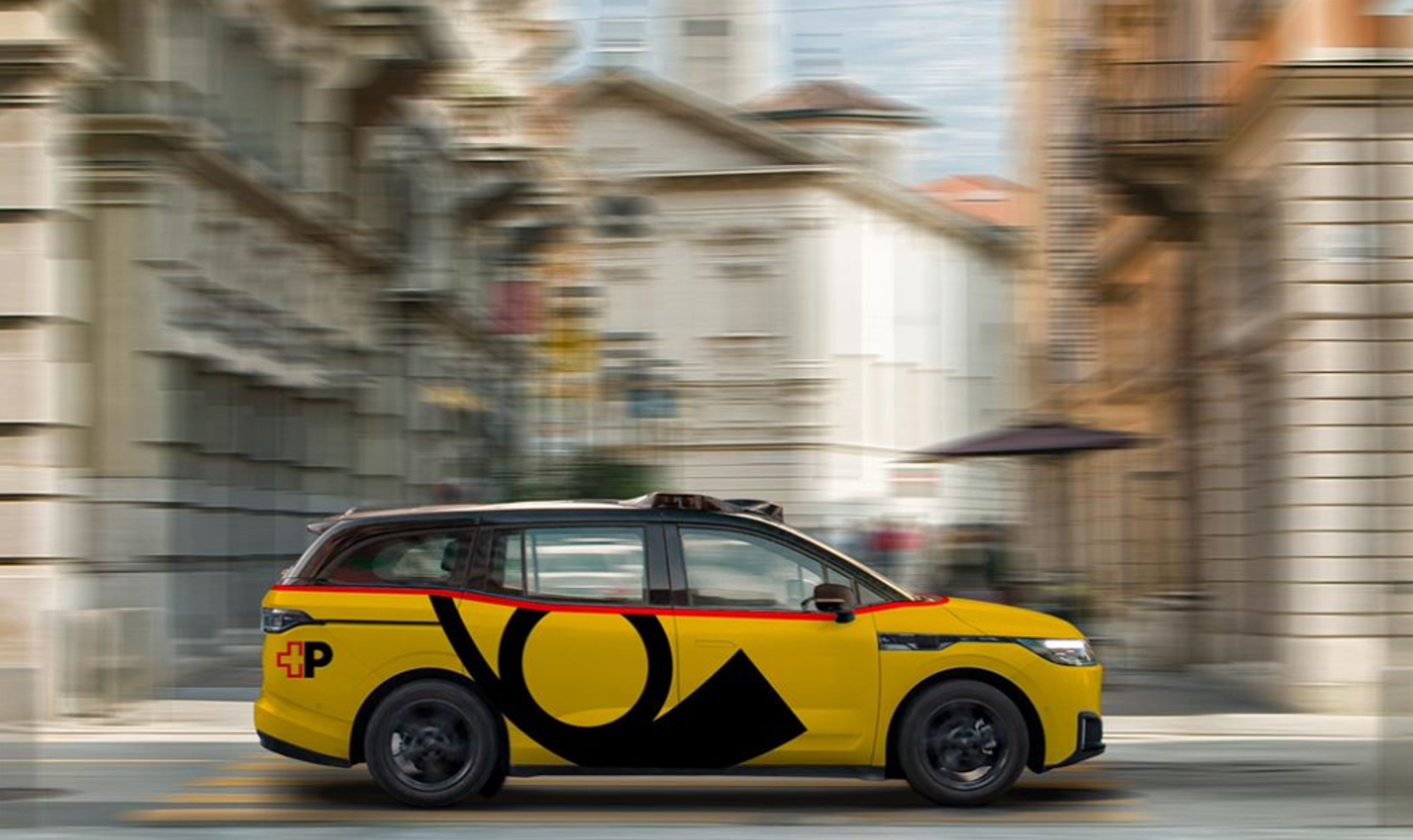 A yellow vehicle with a black logo driving through a street with historic buildings in the background.
