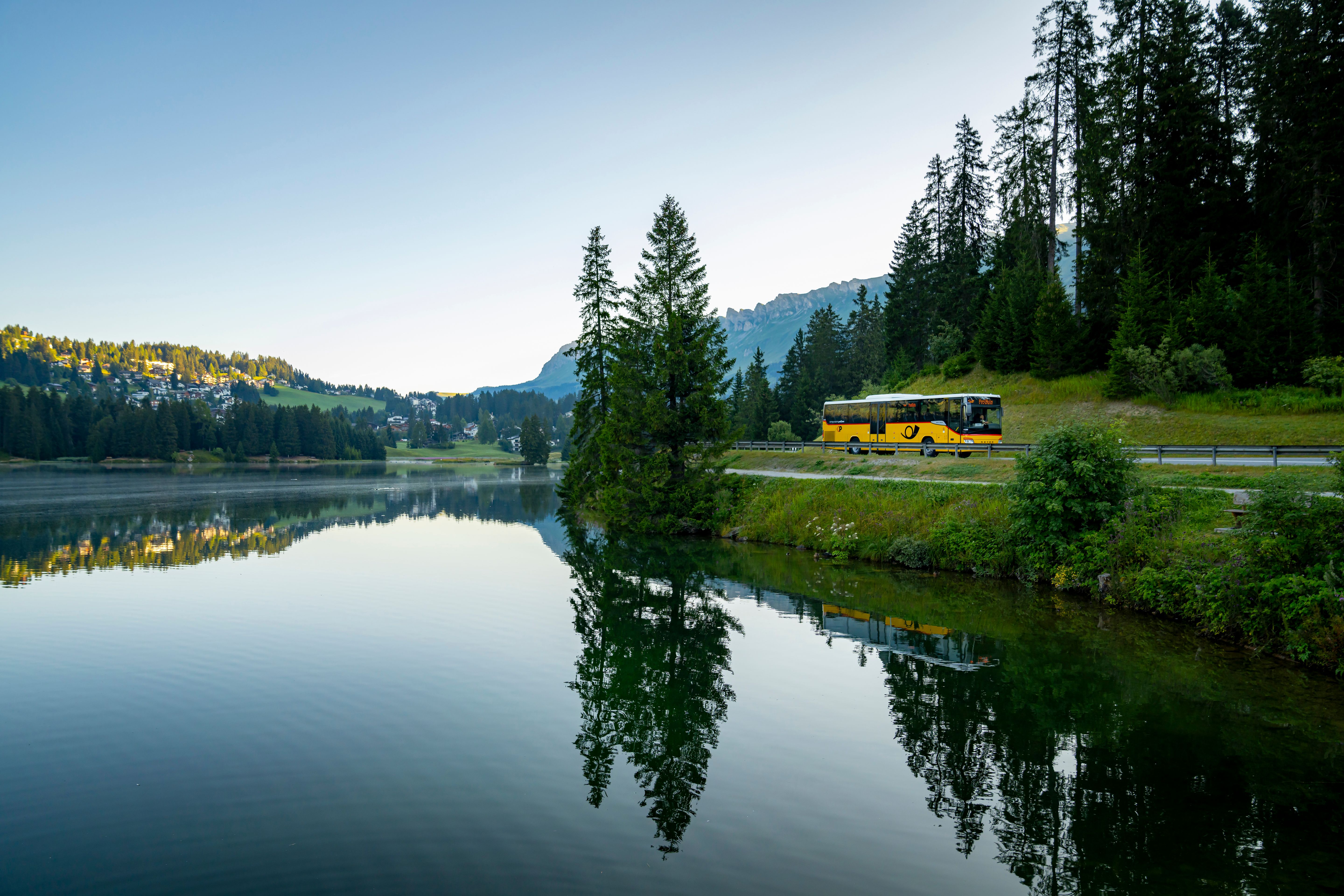 A yellow bus travels along the lake, surrounded by trees and mountains.