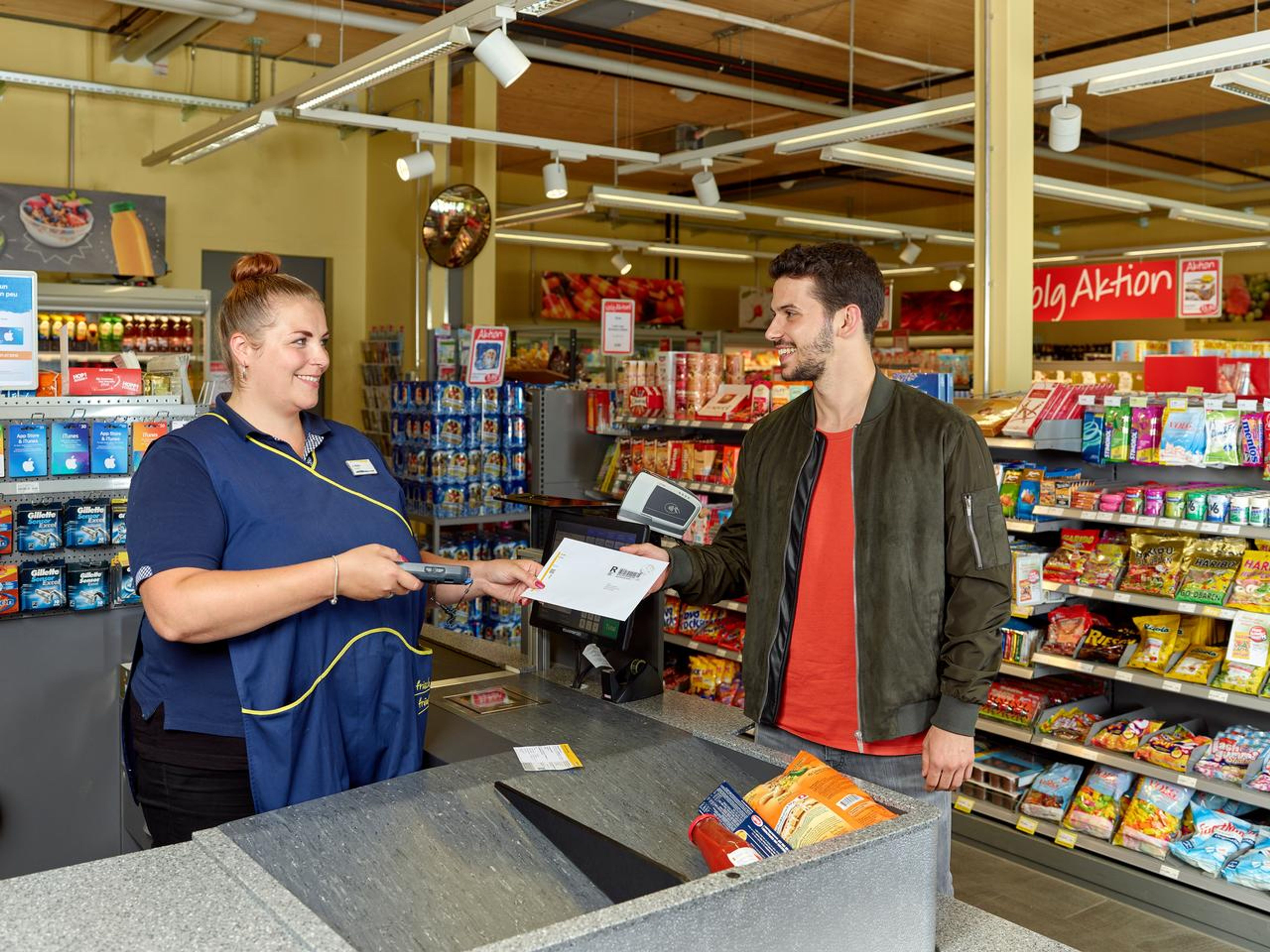 Service postal à la caisse d’un commerce de détail. Une personne remet une enveloppe à la caissière qui tient un lecteur. Sur le comptoir, des snacks ; à l’arrière-plan, des rayons avec des produits alimentaires et une affiche “Aktion”.