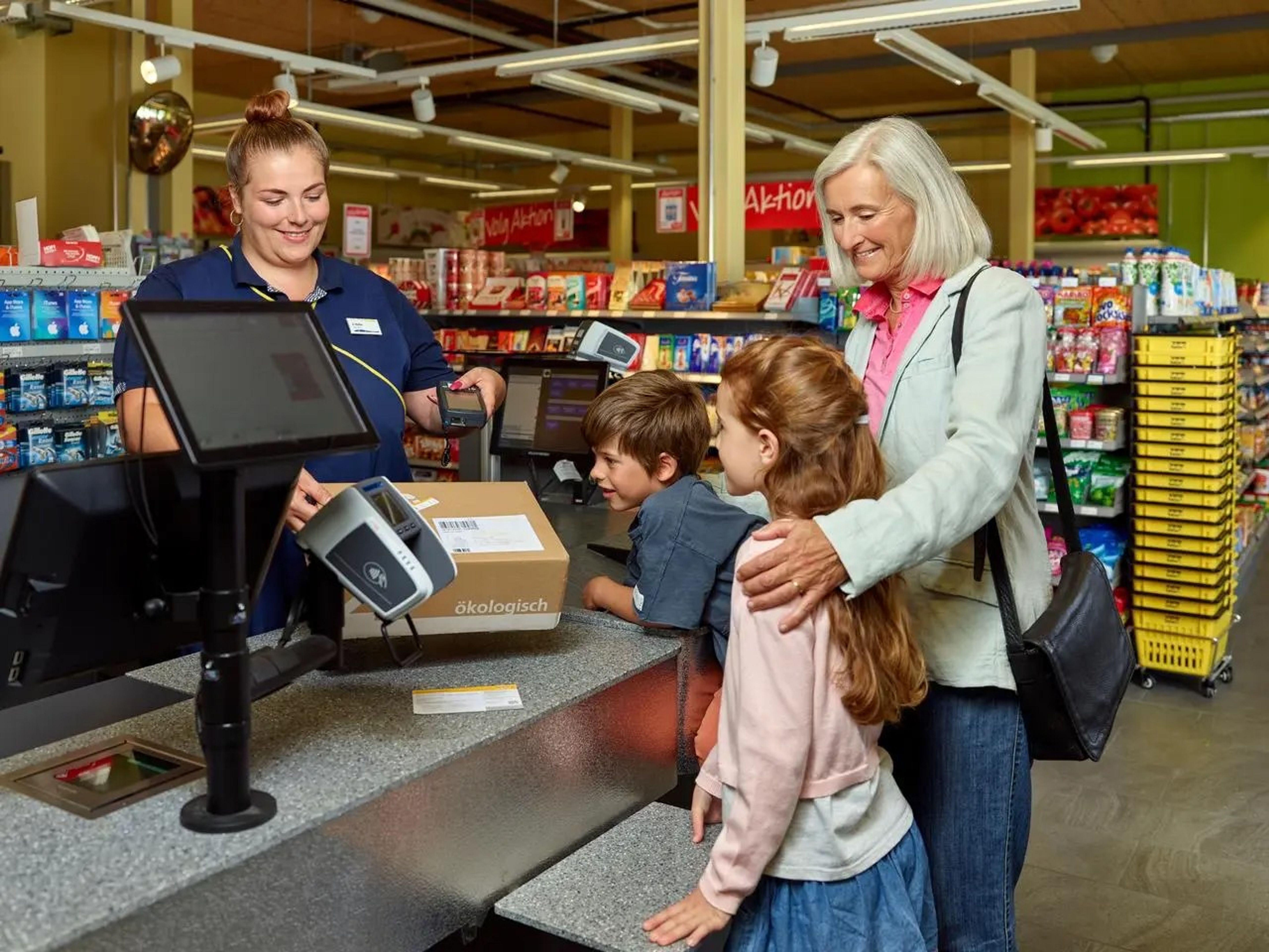 Service postal à la caisse d’un commerce de détail. Sur le comptoir se trouve un colis portant la mention “ökologisch”. Une personne est avec deux enfants au guichet, tandis que la caissière tient un lecteur. À l’arrière-plan, des rayons avec des produits alimentaires et une affiche “Aktion”.