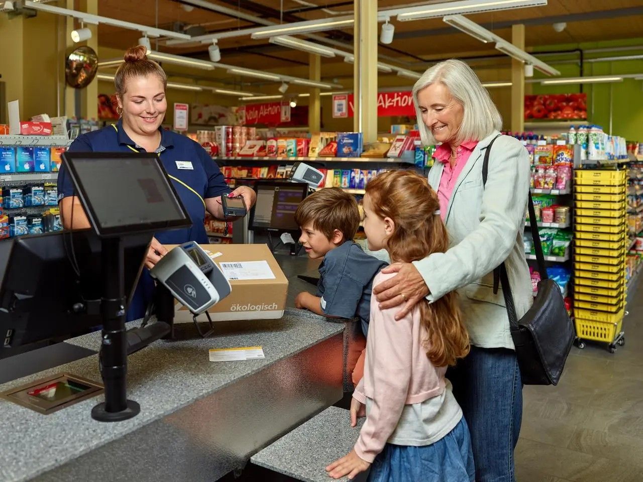 Service postal à la caisse d’un commerce de détail. Sur le comptoir se trouve un colis portant la mention “ökologisch”. Une personne est avec deux enfants au guichet, tandis que la caissière tient un lecteur. À l’arrière-plan, des rayons avec des produits alimentaires et une affiche “Aktion”.