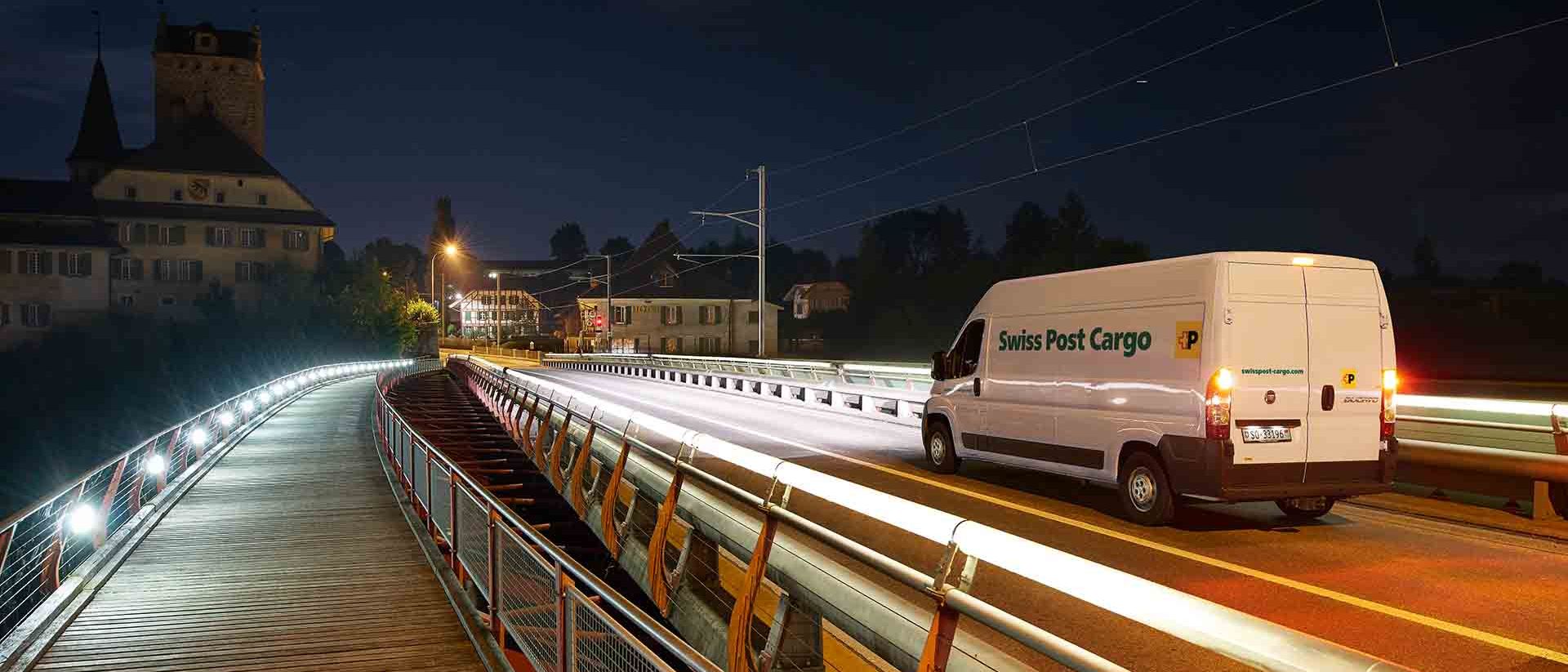 A Swiss Post Cargo delivery vehicle drives over a bridge at night with an express delivery.