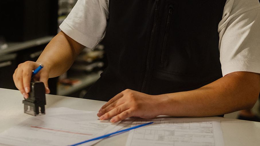 Un agent des douanes tamponne des documents à un guichet, avec un bureau et plusieurs personnes en arrière-plan.