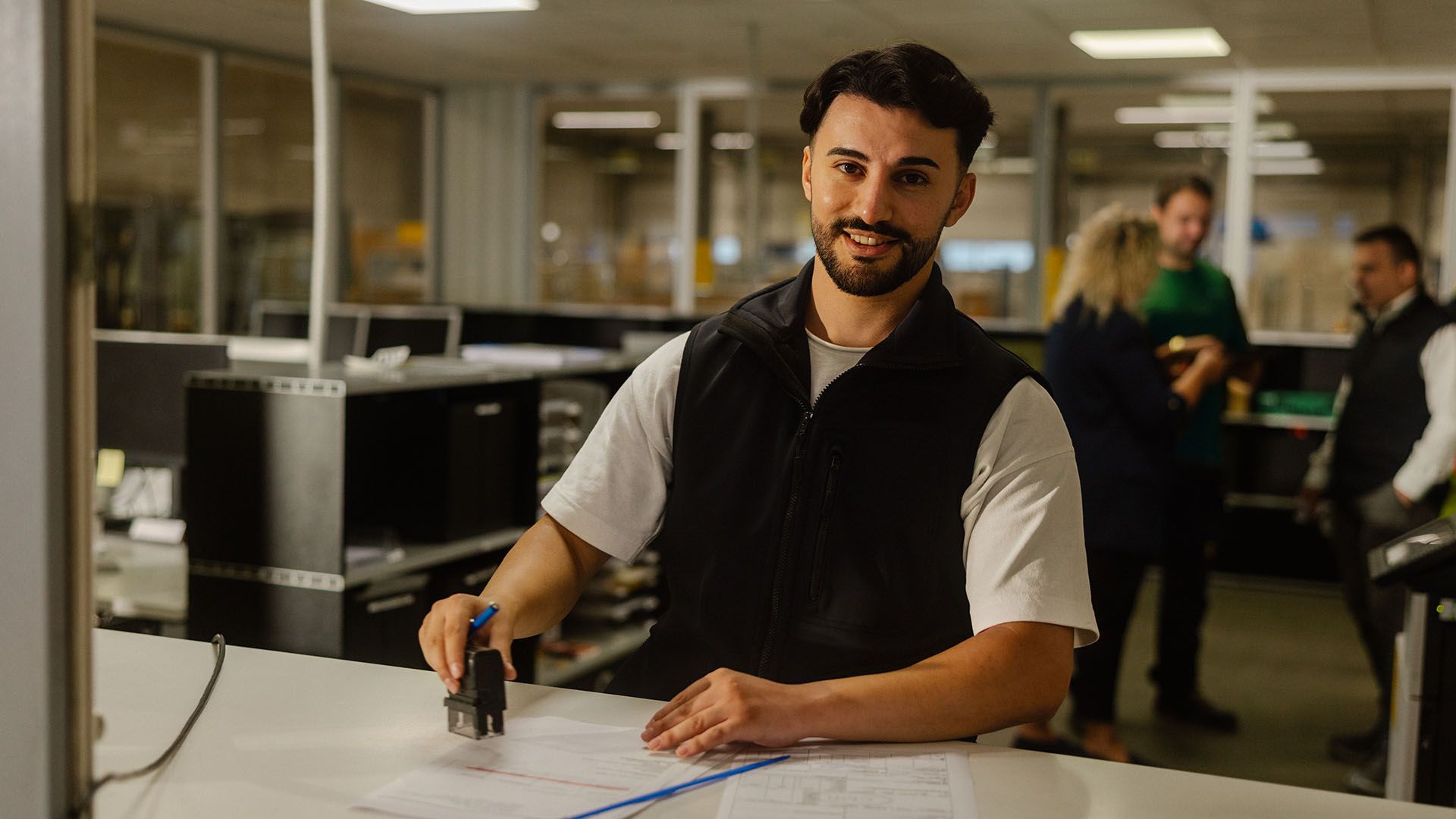 Ein Zollbeamter stempelt Dokumente an einem Schalter, im Hintergrund ein Büro mit mehreren Personen.