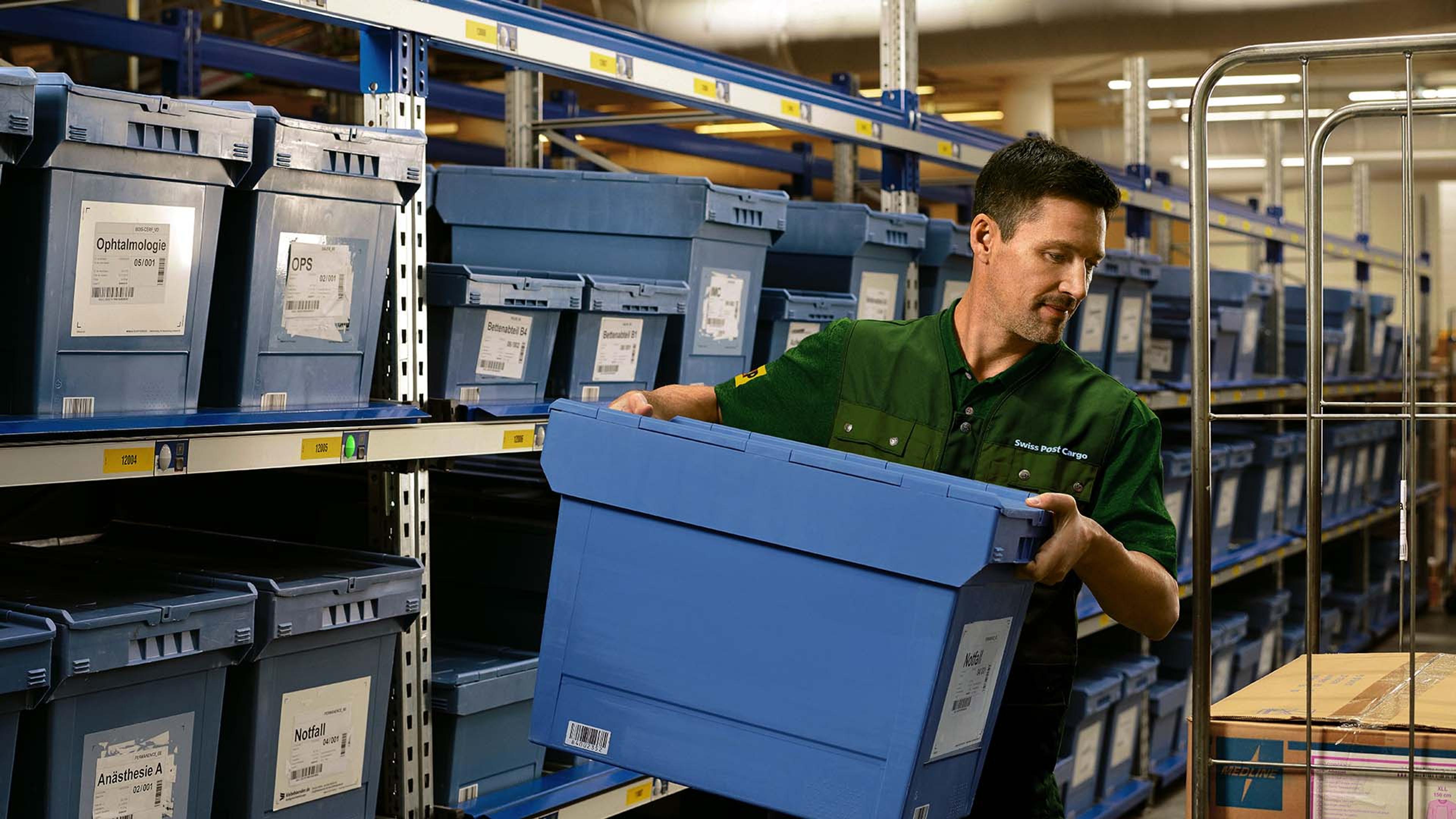 A warehouse worker loads a box with pharmaceutical goods from a shelf onto a trolley.