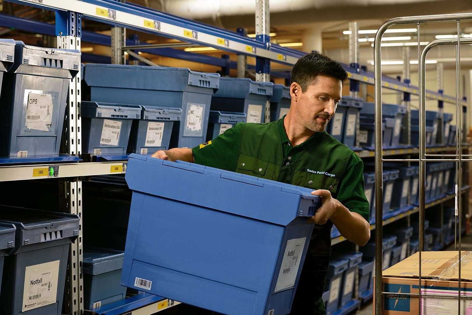 A warehouse worker loads a box with pharmaceutical goods from a shelf onto a trolley.