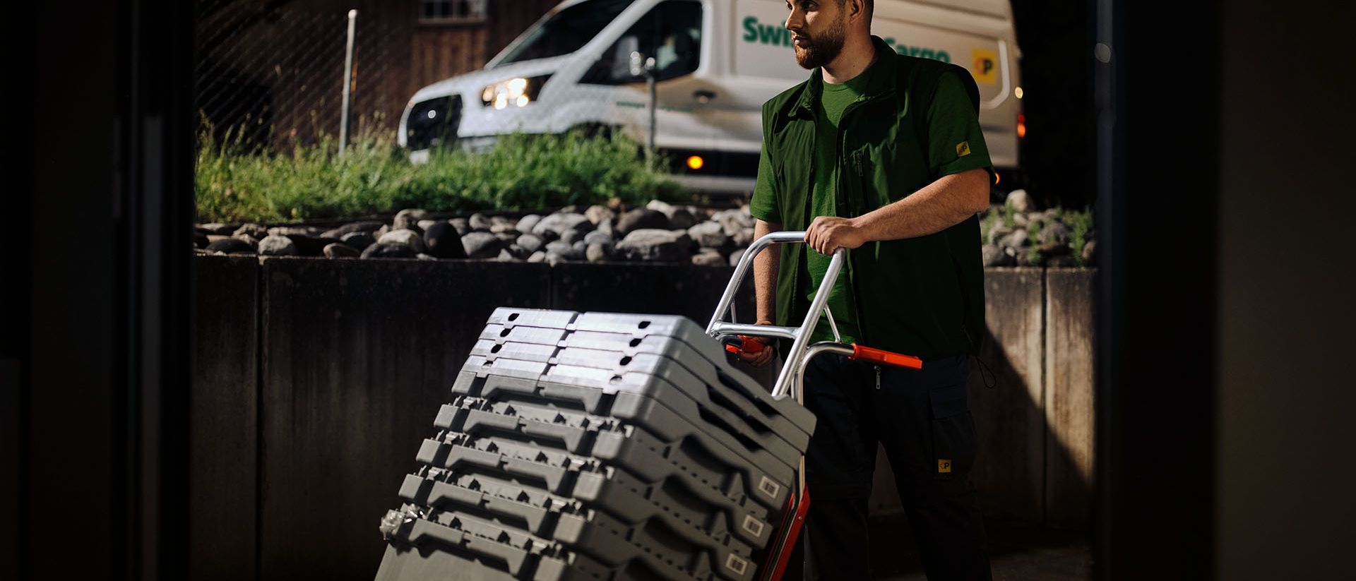 A Swiss Post Cargo night courier delivers medical goods with a hand trolley, with a delivery vehicle in the background.