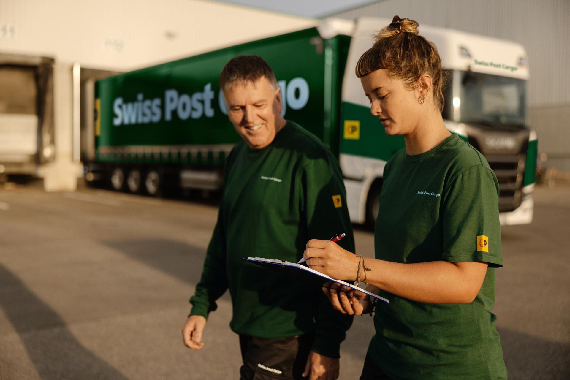 Zwei Swiss Post Cargo Mitarbeitende gehen an einem Swiss Post Cargo LKW vorbei, der an einer Laderampe steht.