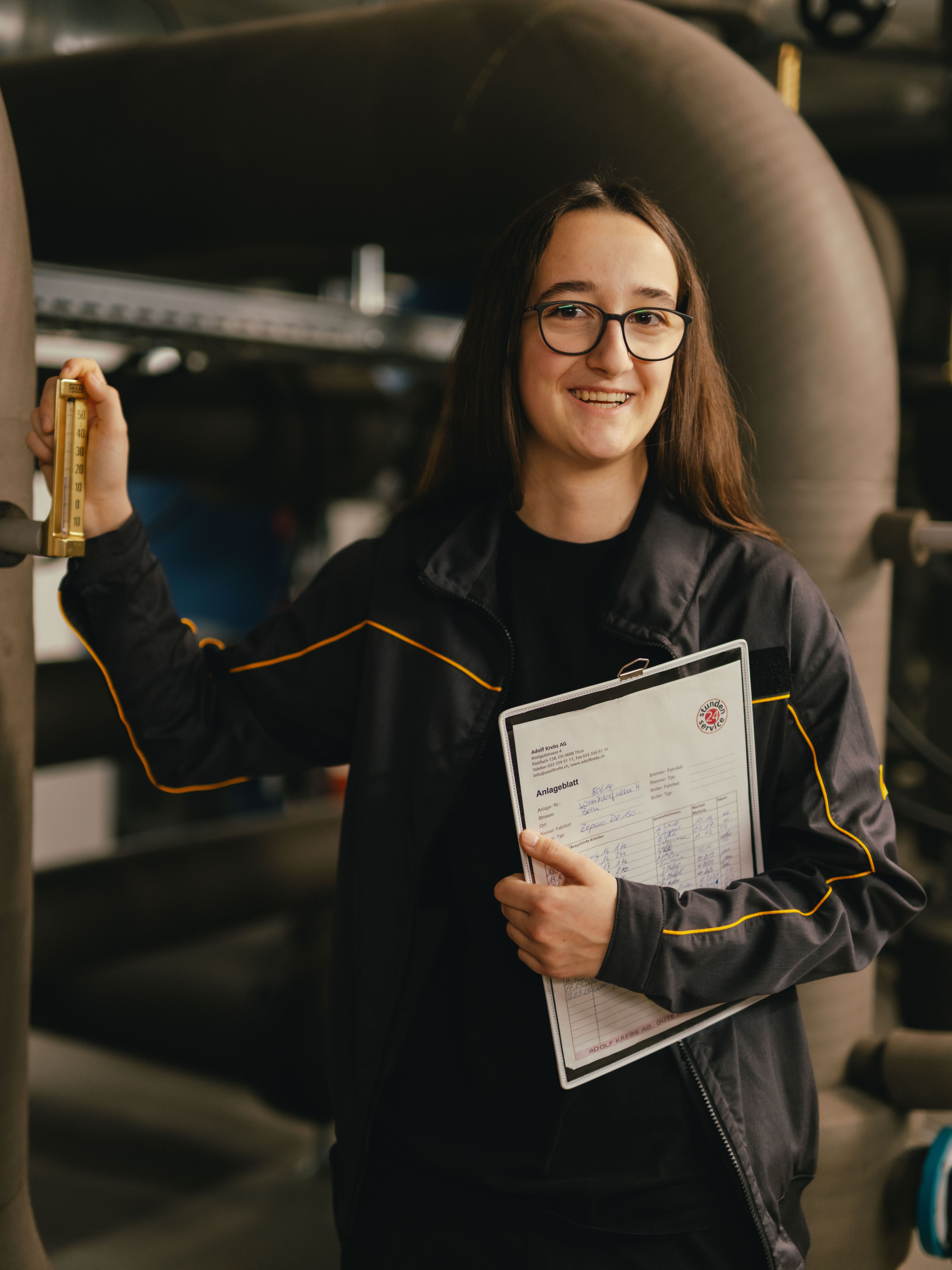A person is standing between large pipes in a technical facility and holding a lever on one of the pipes. In their other hand, the person is holding technical documents or a plan. They are wearing dark work clothing with light contrast seams. In the background, other pipelines, valves and metal structures in an industrial environment can be seen.