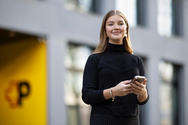 Eine Person steht im Freien vor einem modernen Gebäude und hält ein Smartphone in beiden Händen. Die Person trägt einen dunklen langen Pullover und ein Armband. Im Hintergrund ist eine gelbe Wand mit dem Post-Logo zu sehen.