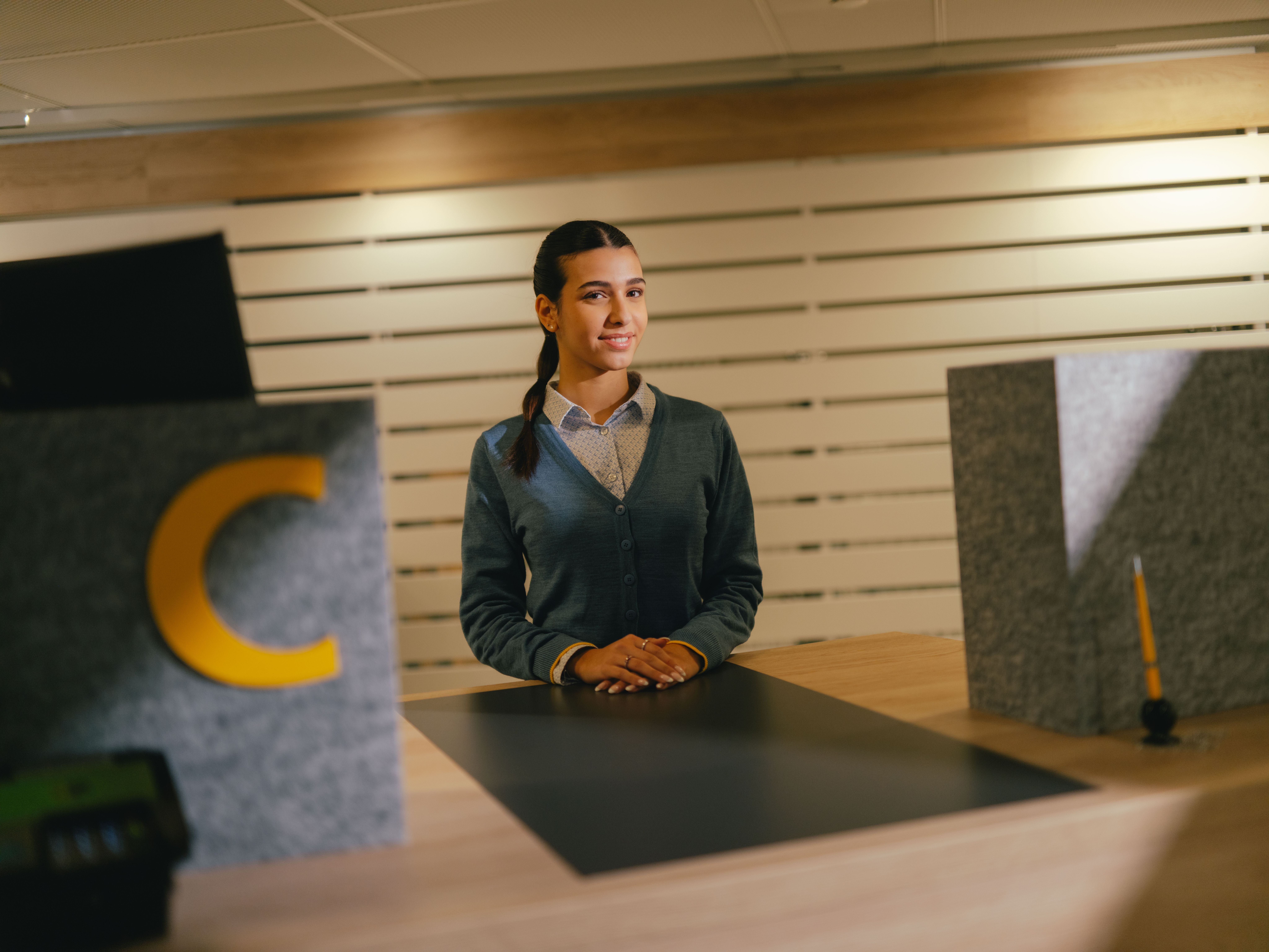 A person stands behind a counter in a reception or service area. In front of them is a dark work surface, framed by two grey, structured elements, one of which bears a large yellow letter “C”. The background consists of bright slats of wall arranged horizontally. The person is wearing a light shirt and dark cardigan and has put their hands down in front of their body.