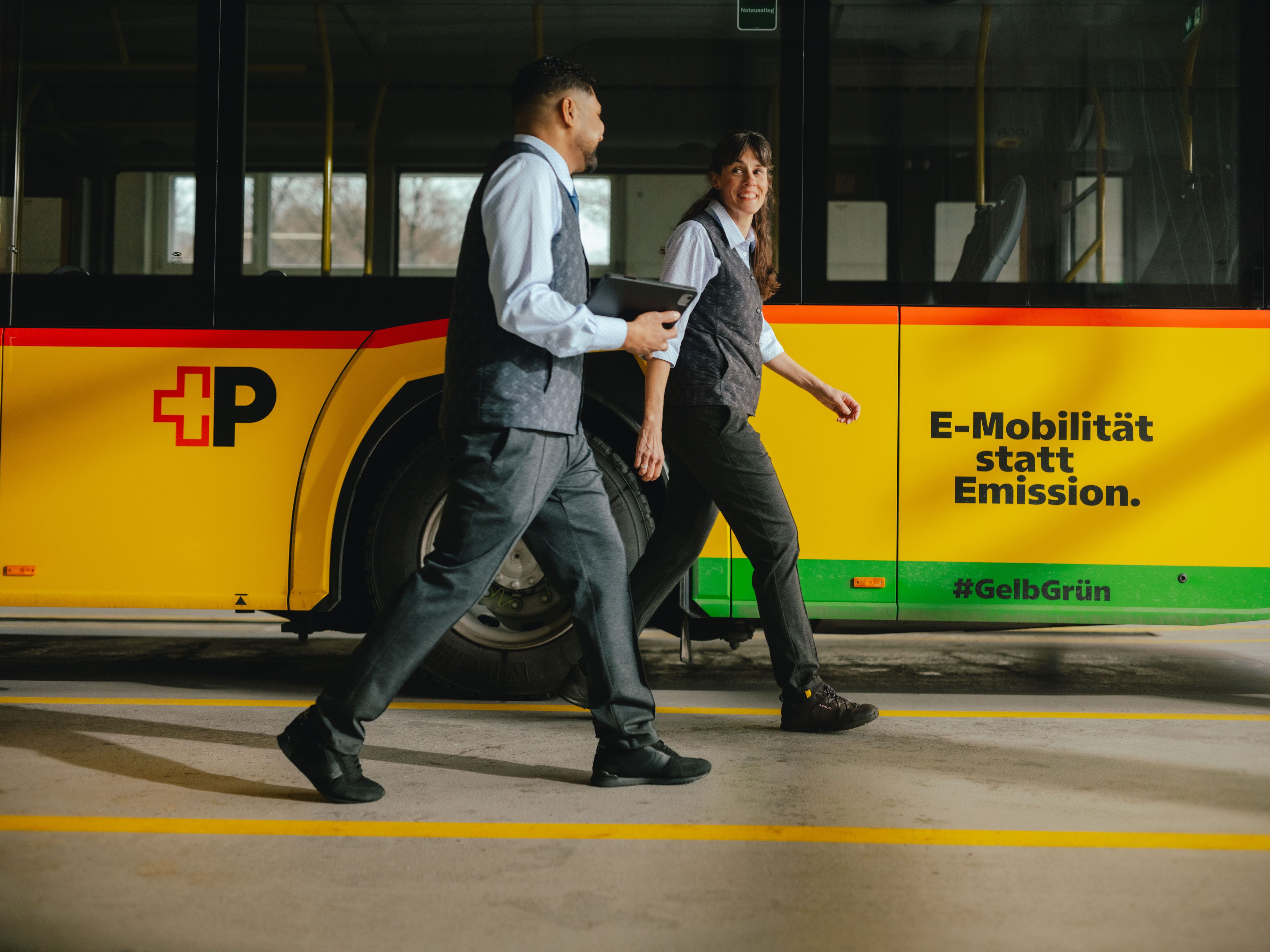 Deux personnes marchent côte à côte dans un hall, le long d’un bus jaune et noir. L’une des personnes tient une tablette à la main. Sur le côté du bus, on peut lire « E‑Mobilité statt Emission. » ainsi que des hashtags et des bandes colorées. L’environnement ressemble à un dépôt ou un atelier de maintenance.