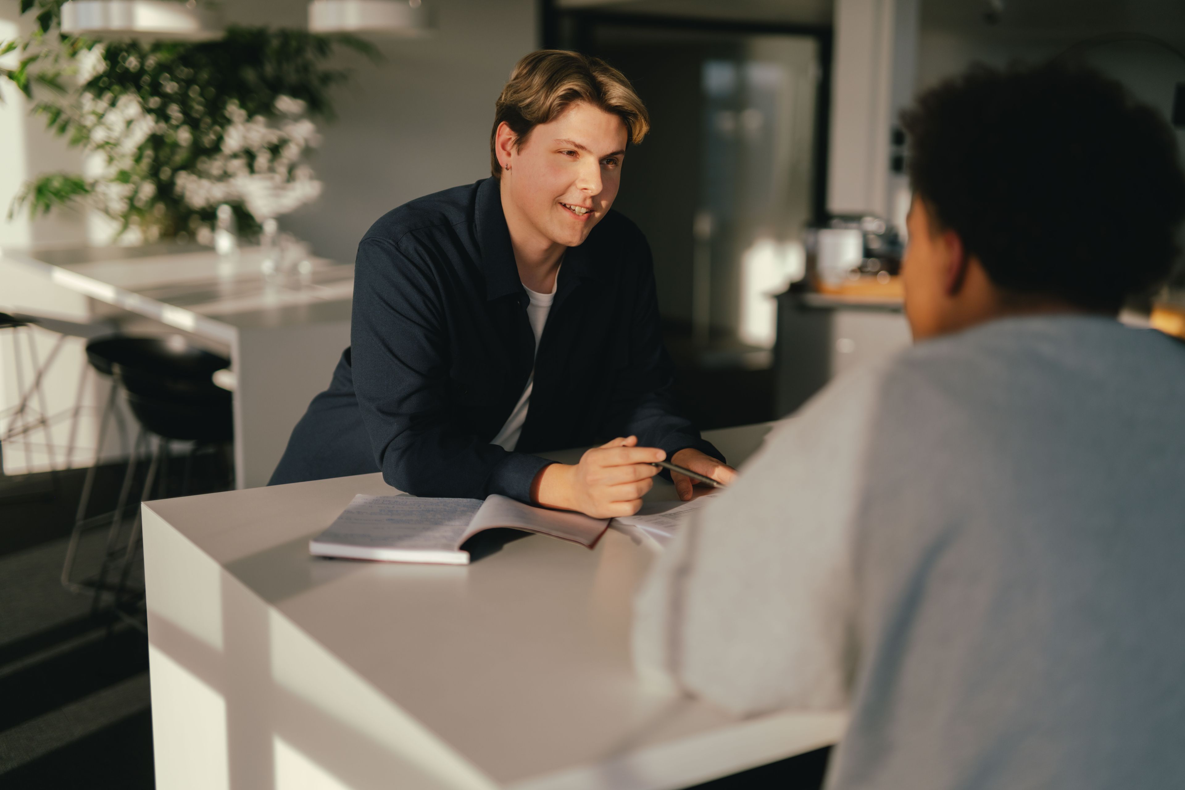 Deux personnes se font face à une table dans un intérieur moderne. L’une des personnes tient un stylo sur un bloc-notes ouvert, comme si quelque chose était expliqué ou discuté. D’autres documents sont posés sur la table et, en arrière-plan, on distingue des plantes, des meubles et de grandes fenêtres. La lumière pénètre latéralement dans la pièce et produit des ombres douces.