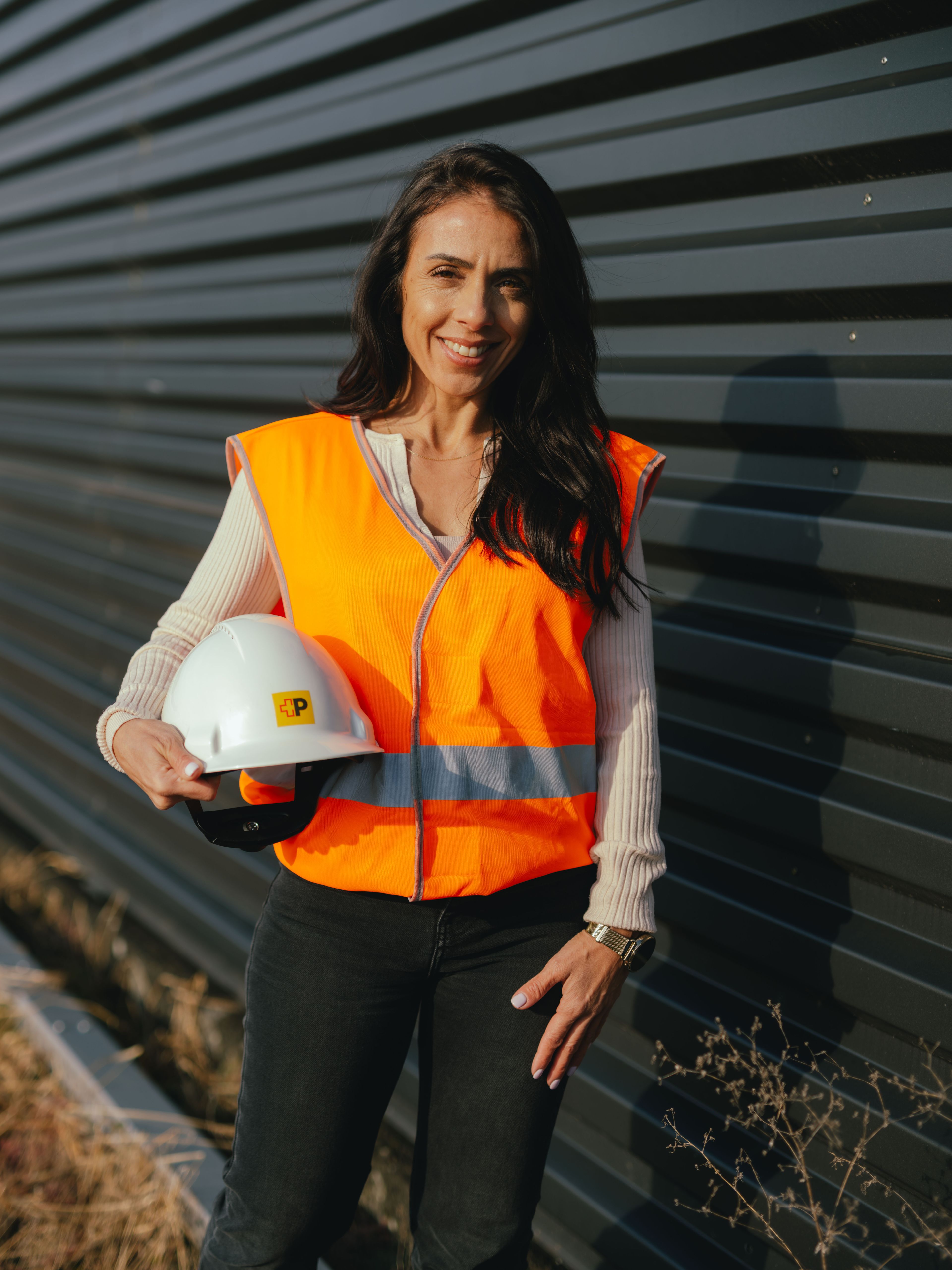 A person is standing outdoors next to a long, dark metal wall. The person is wearing a bright orange safety vest and holding a white protective helmet. In the foreground, dry grasses are visible, and the sunlight casts strong shadows.