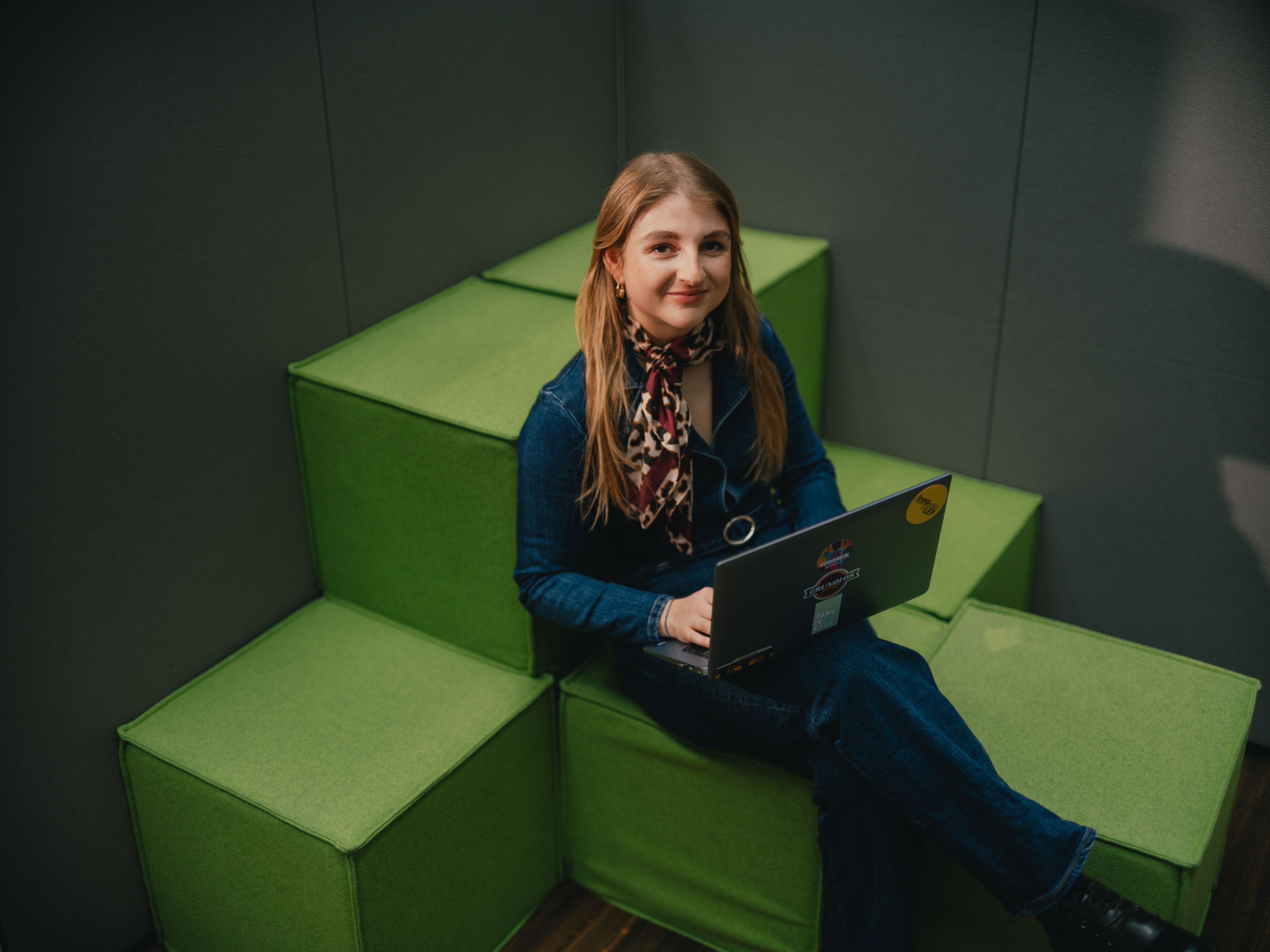 Eine Person sitzt auf grünen, stufenförmig angeordneten Polsterelementen und arbeitet mit einem aufgeklappten Laptop auf dem Schoss. Die Person trägt dunkle Kleidung und ein gemustertes Tuch um den Hals. Der Raum wirkt gedämpft beleuchtet, und die Wände um die Sitzfläche sind dunkel gehalten.