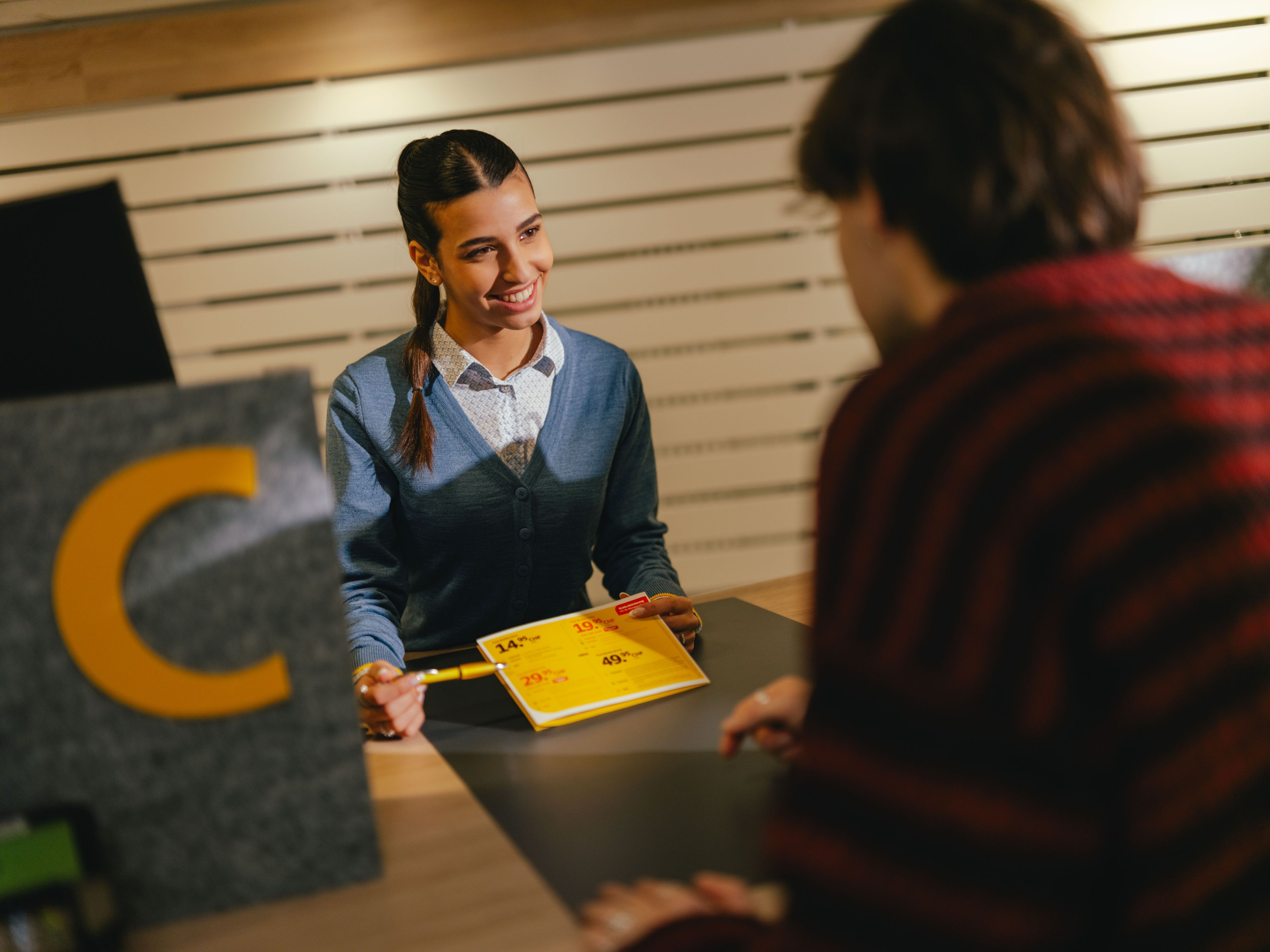 One person sits at a counter and hands a yellow form to another person over the counter. The counter area is marked with a grey element showing a large yellow letter “C”. The surroundings look like a reception or service area with light wooden slats in the background.
