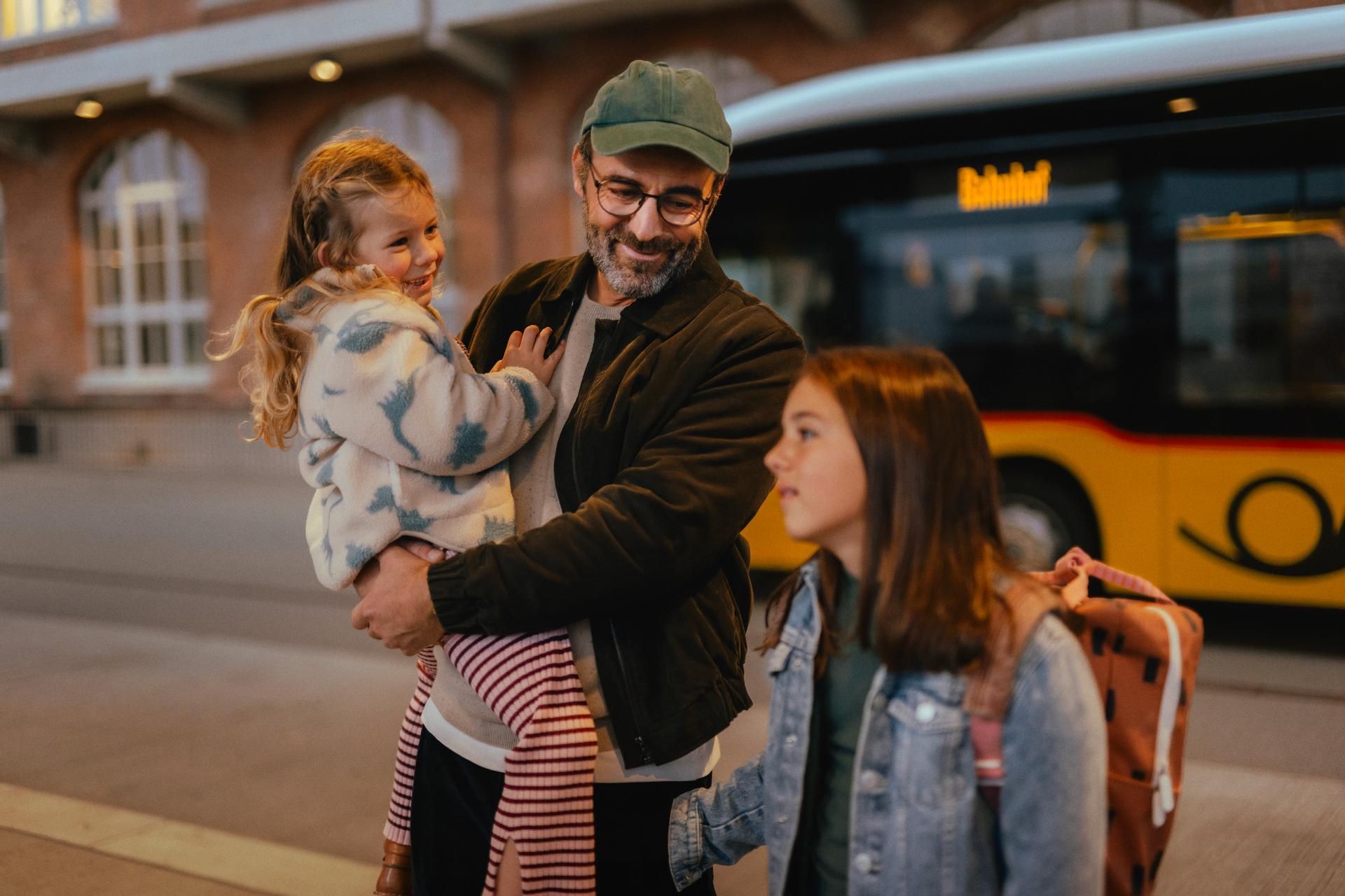 Tre persone si trovano a una fermata dell’autobus davanti a un autobus giallo. Una persona tiene un bambino in braccio, mentre un’altra persona con uno zaino è accanto. Sullo sfondo si vede un edificio in mattoni con finestre illuminate.