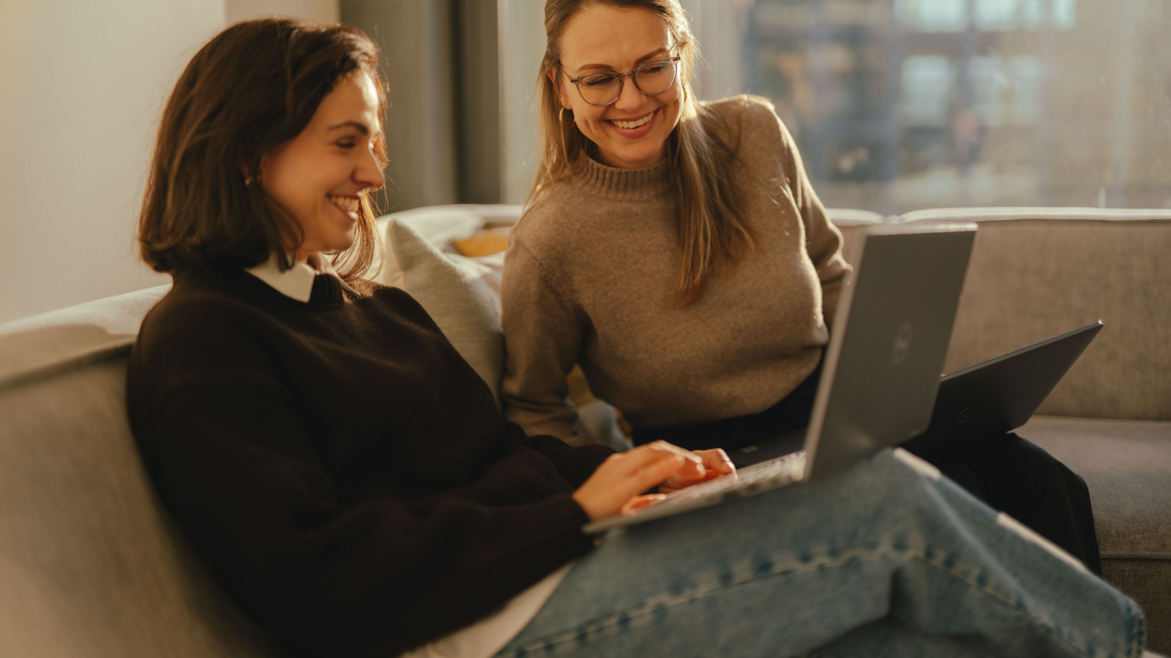 Two individuals are seated on a sofa in a modern indoor space, each working on an open laptop. The individuals are sitting close together, with one of the laptops resting on a person’s lap. Large windows in the background reveal urban buildings and allow warm light into the room.