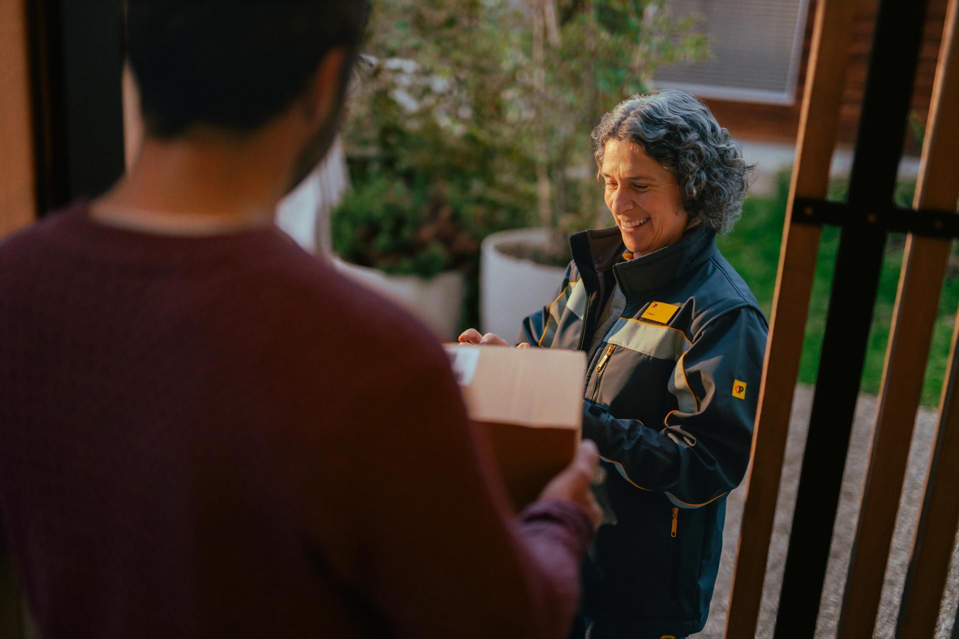 Una persona con una divisa da consegna porge un pacco a un’altra persona in primo piano. Lo scambio avviene davanti a una porta d’ingresso, con piante ed elementi in legno sullo sfondo.