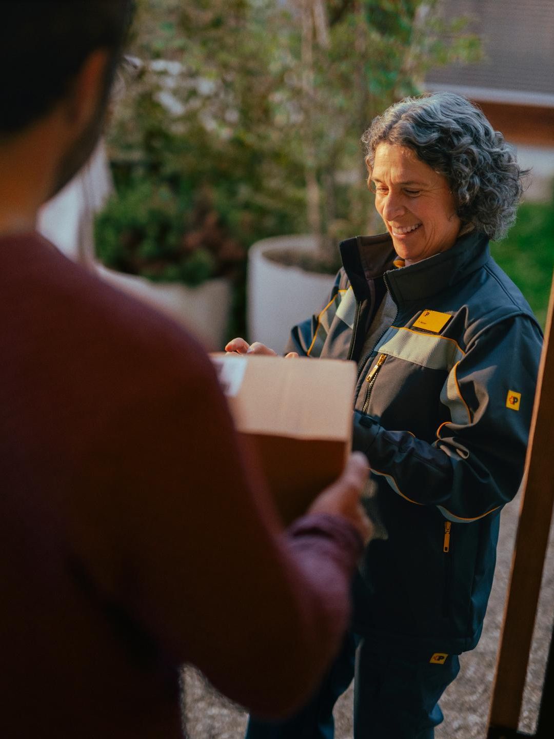A person in a delivery uniform hands a package to another person in the foreground. The exchange takes place at a front door, with plants and a wooden structure visible in the background.