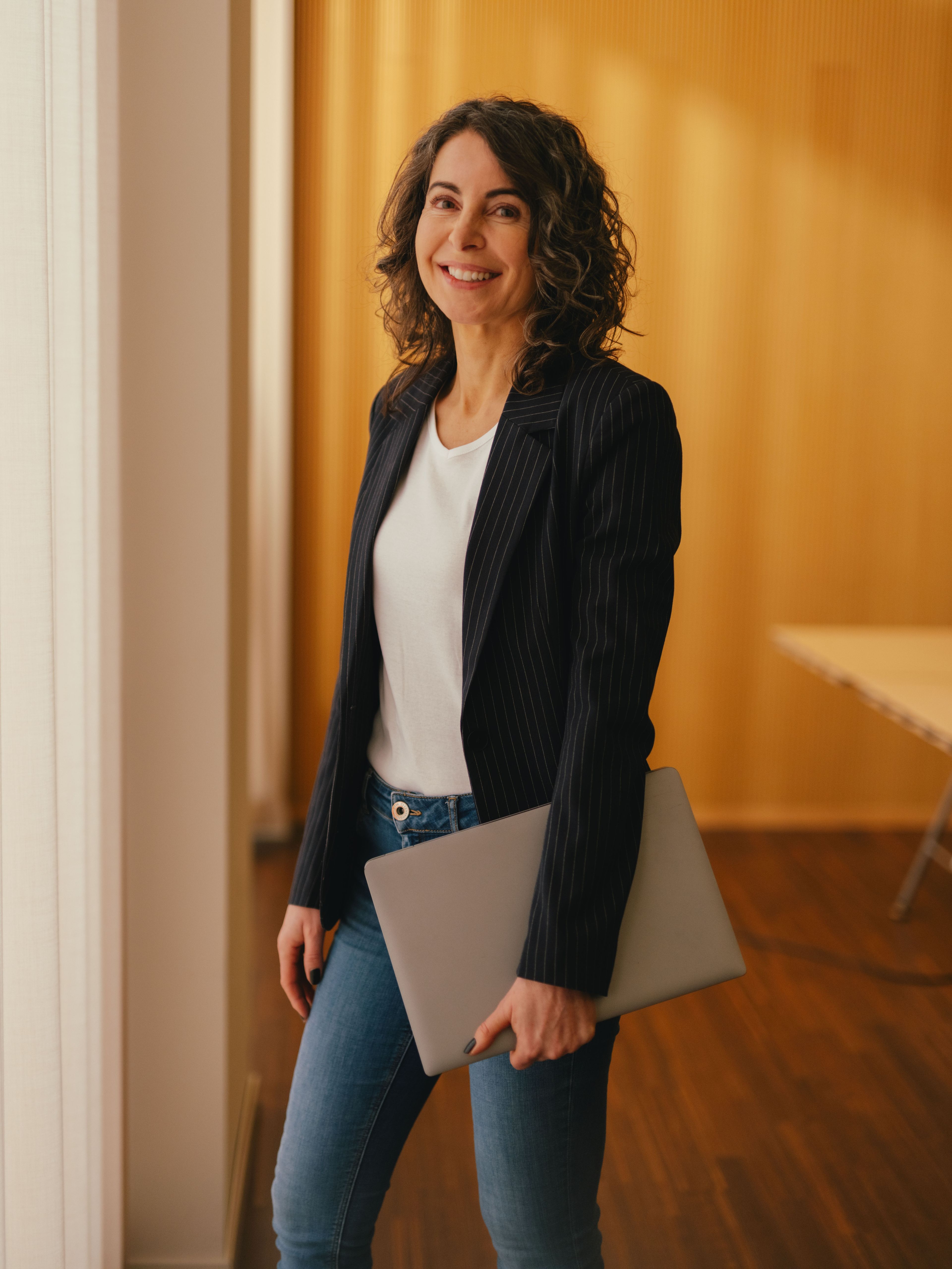 A person stands in a room with light wooden walls, holding a closed laptop at their side. The person is wearing a dark jacket, a light shirt, and jeans. Natural light enters through a large window, and a table with a monitor is visible in the background.