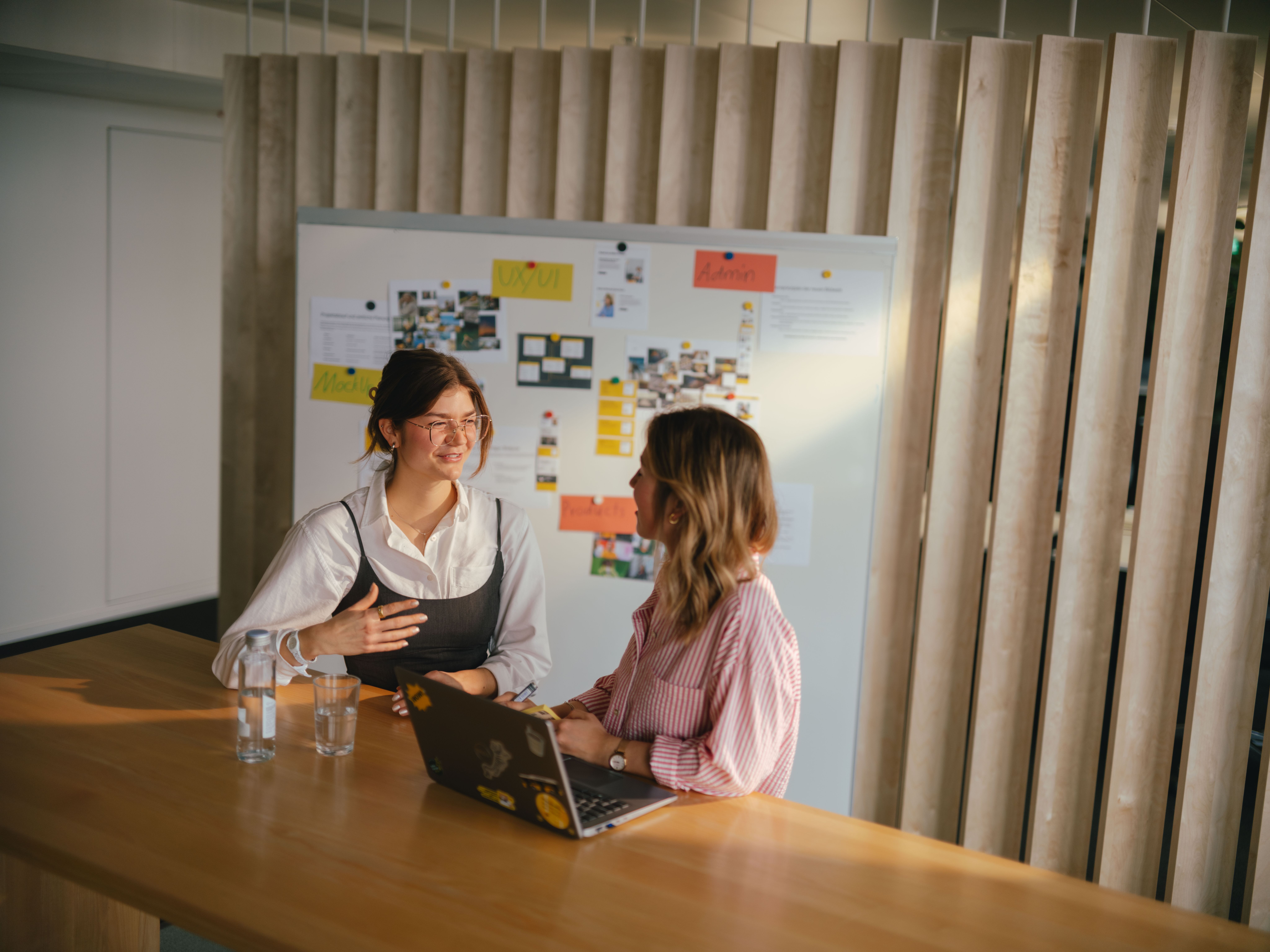 Due persone sono sedute a un lungo tavolo di legno in un ambiente interno moderno. Davanti a loro c’è un computer portatile aperto con diversi adesivi e due bicchieri d’acqua. Sullo sfondo si vede una grande bacheca con numerosi post‑it, foto e cartoncini colorati. Dietro la bacheca sono presenti pannelli di legno verticali attraverso cui entra una luce calda.