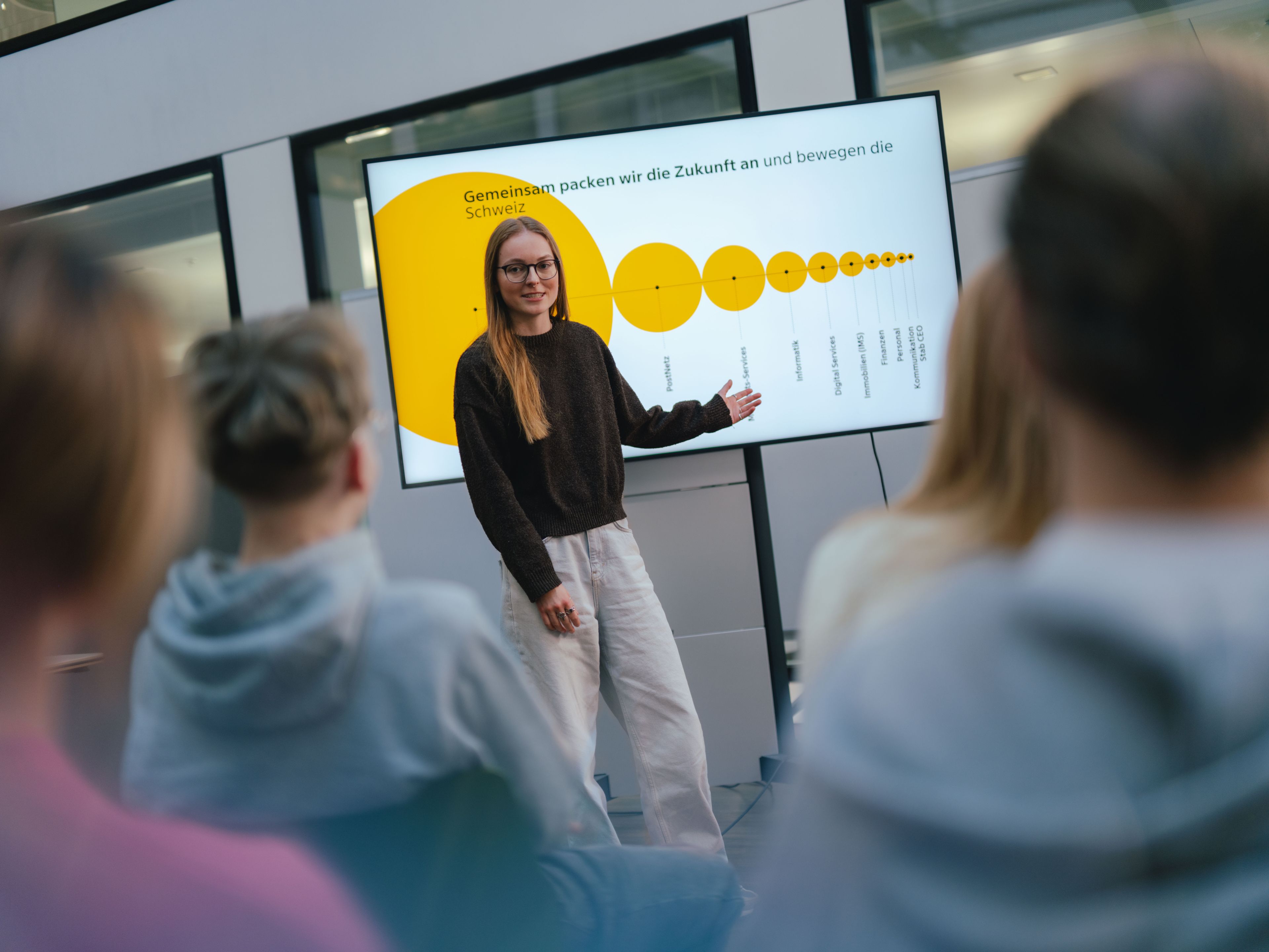 A person stands in front of a large screen and gives a presentation for a seated audience. The screen shows a yellow graph with circles of different sizes and dots. The room has large windows, and several people are visible from behind in the foreground.
