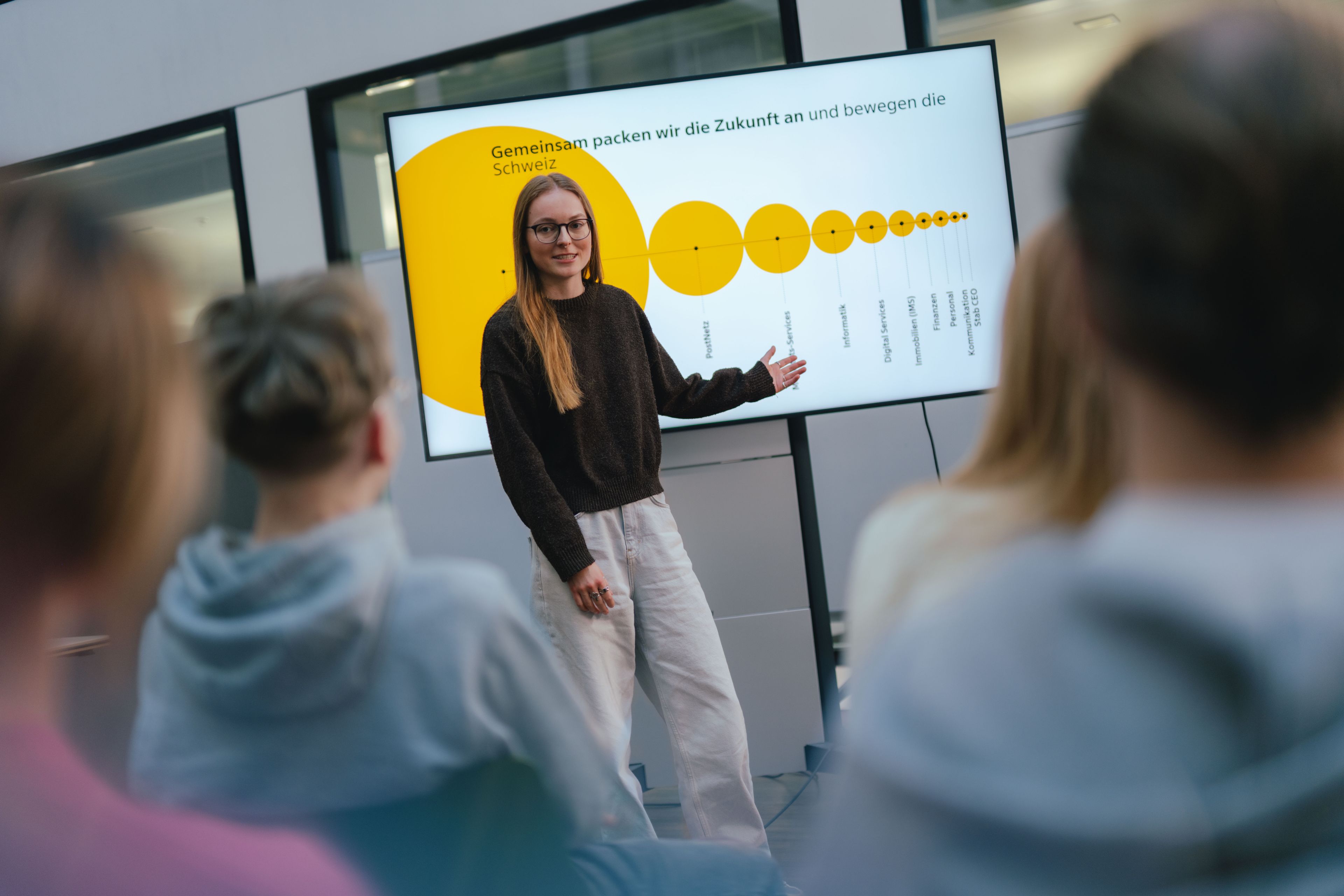 Une personne se tient devant un grand écran et fait une présentation pour un public assis. L’écran affiche un graphique jaune avec des cercles de différentes tailles et des points marqués. La salle est dotée de grandes fenêtres et plusieurs personnes sont visibles de l’arrière-plan.