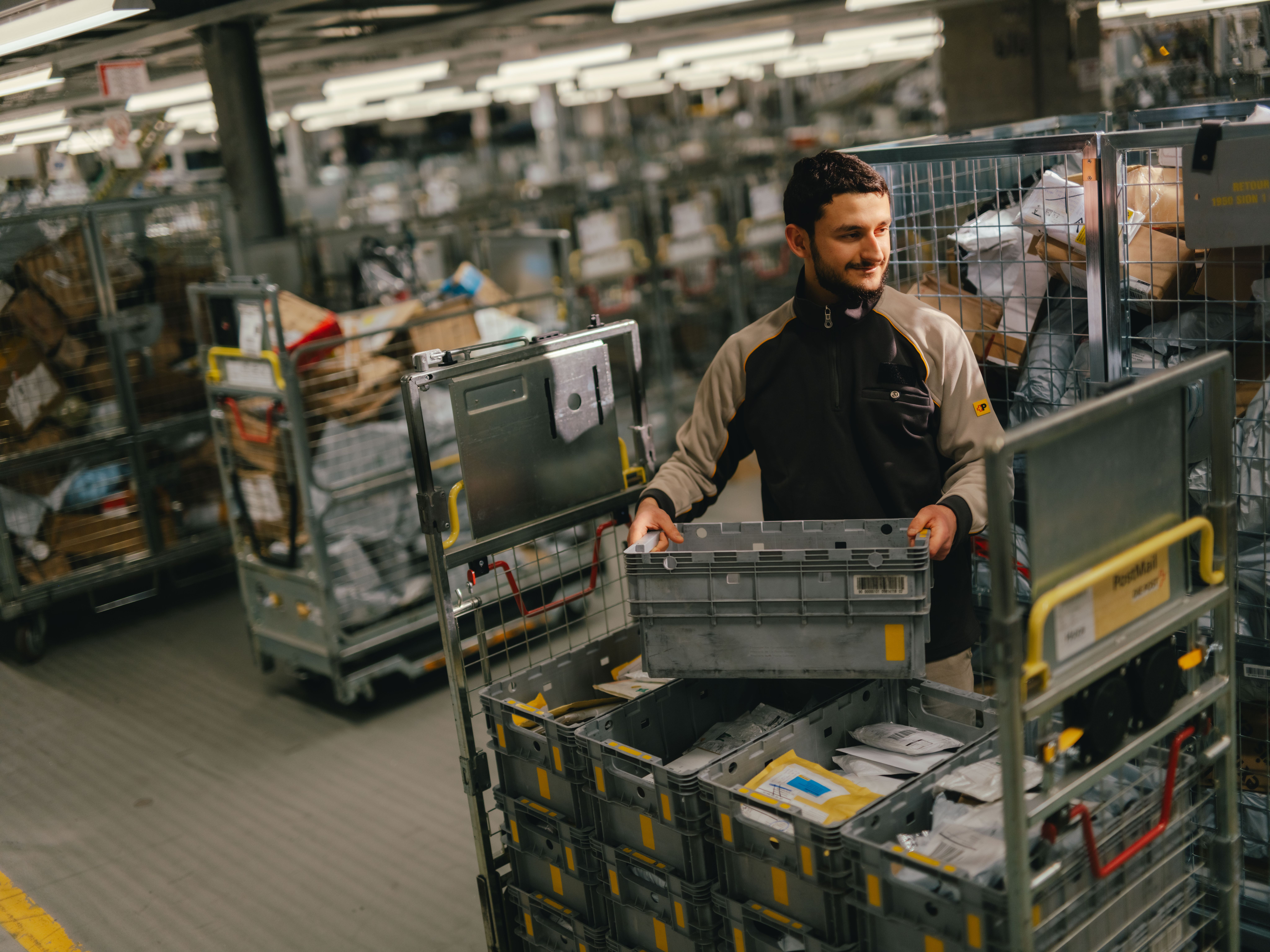 Une personne se tient dans un grand hall de tri et soulève une caisse de transport grise. La personne porte une veste de travail sombre avec des bandes contrastées claires. Tout autour se trouvent de nombreux chariots métalliques remplis de colis, d’envois et d’autres bacs. L’espace est bien éclairé et paraît très chargé en matériel de tri et de stockage.