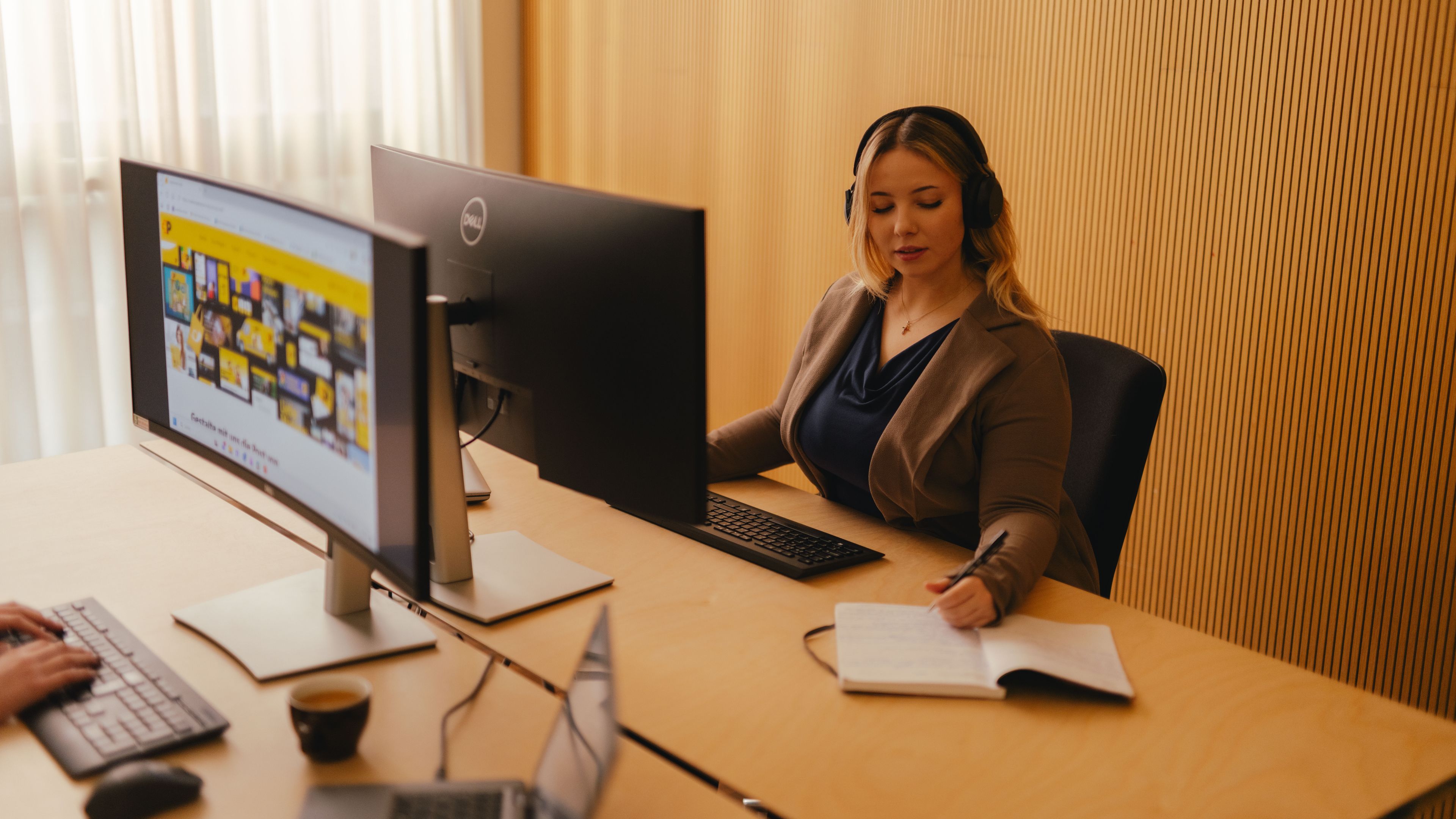 Eine Person sitzt an einem Schreibtisch in einem Büro und arbeitet mit einem grossen Monitor sowie einer Tastatur. Die Person trägt ein Headset und hat ein geöffnetes Notizbuch vor sich. Auf einem zweiten Schreibtisch im Vordergrund steht ein weiterer Laptop. Der Raum hat helle Vorhänge und eine Wand mit vertikalem Muster.