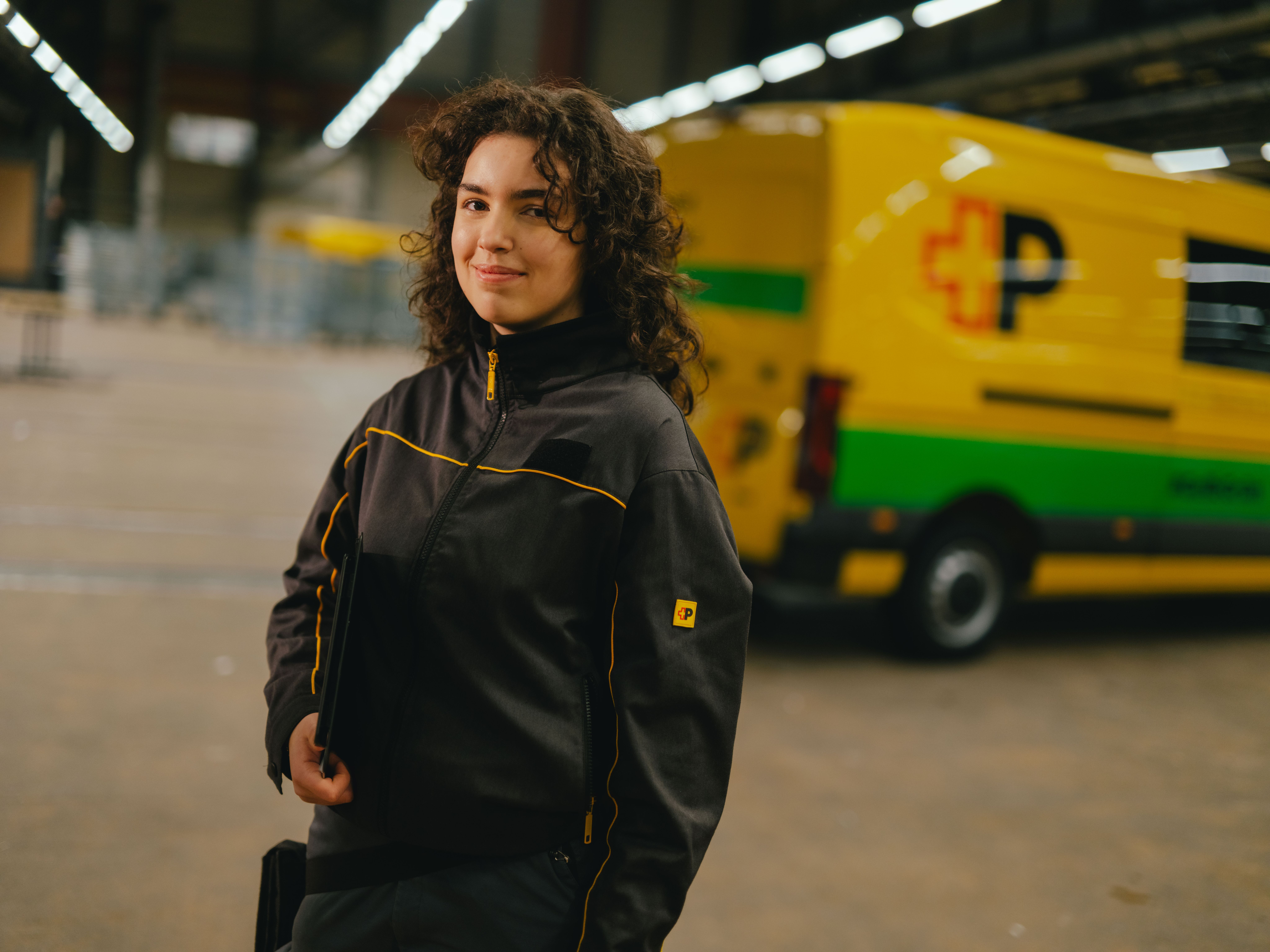 Una persona con abbigliamento da lavoro scuro con dettagli gialli si trova in un grande capannone. La persona tiene un oggetto in mano. Sullo sfondo si vede un veicolo di consegna giallo e verde con scritte nere e rosse e un ampio spazio di magazzino ben illuminato.