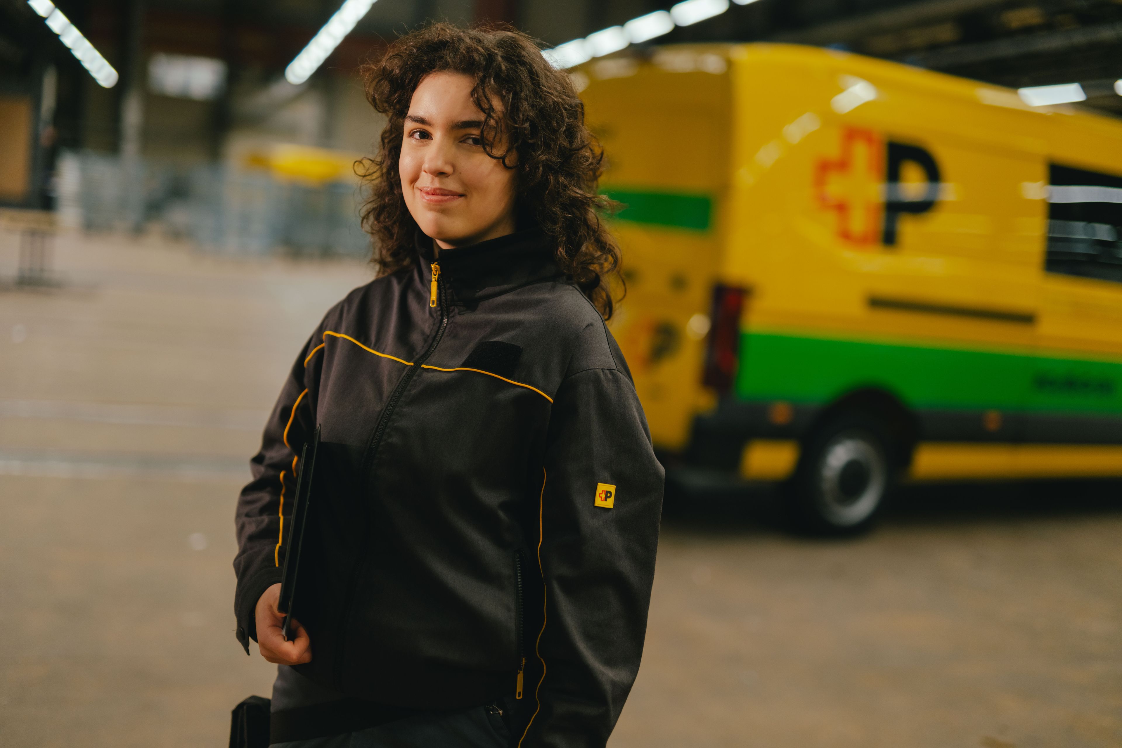 Una persona con abbigliamento da lavoro scuro con dettagli gialli si trova in un grande capannone. La persona tiene un oggetto in mano. Sullo sfondo si vede un veicolo di consegna giallo e verde con scritte nere e rosse e un ampio spazio di magazzino ben illuminato.