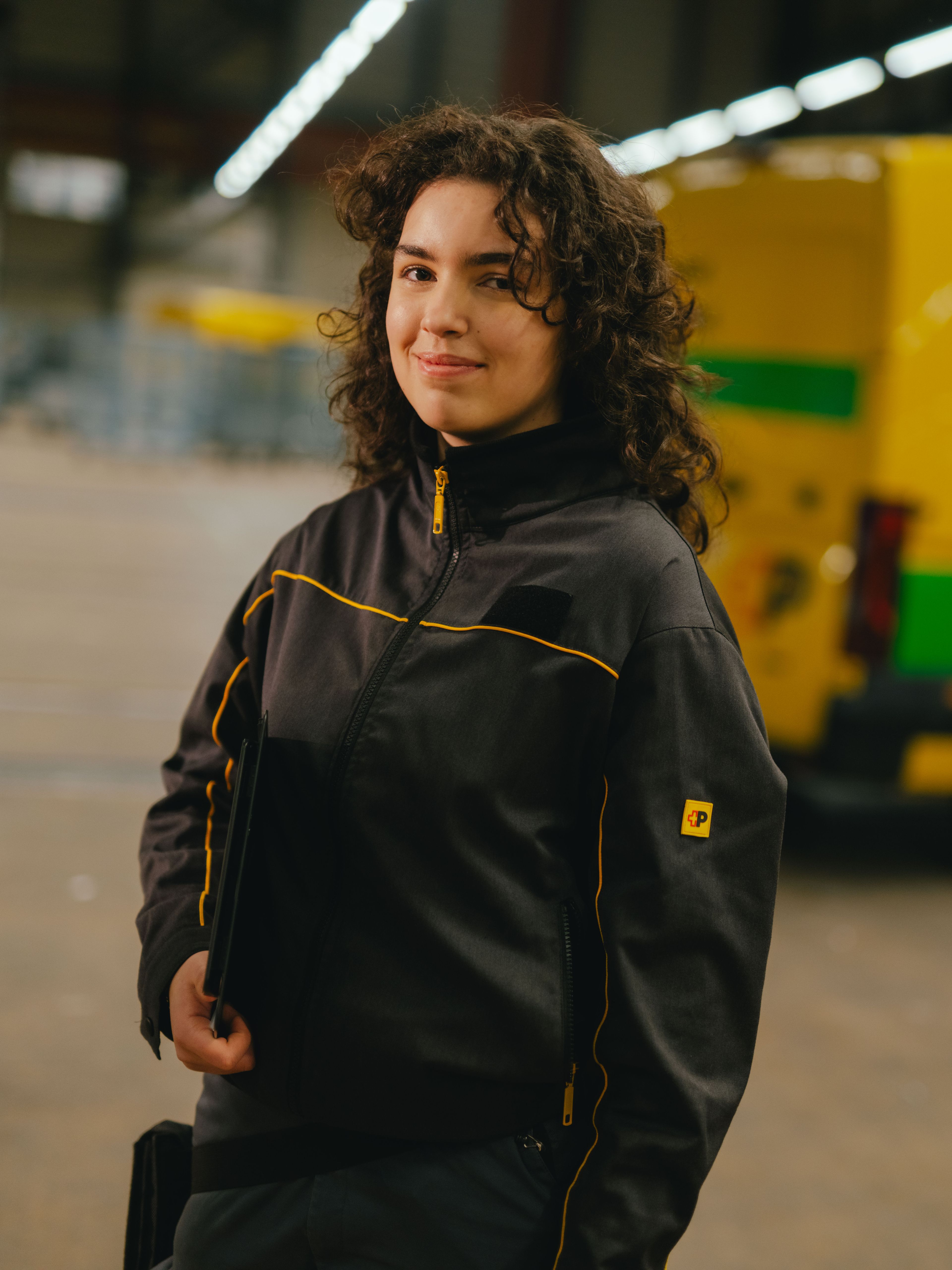 Una persona si trova in un grande padiglione e indossa abiti da lavoro grigio scuro con dettagli gialli. La persona tiene a sé un tablet o un portablocco scuro. Sullo sfondo si vede un ‑veicolo per consegne giallo-verde con scritta nera, nonché strutture industriali e capannoni alti.