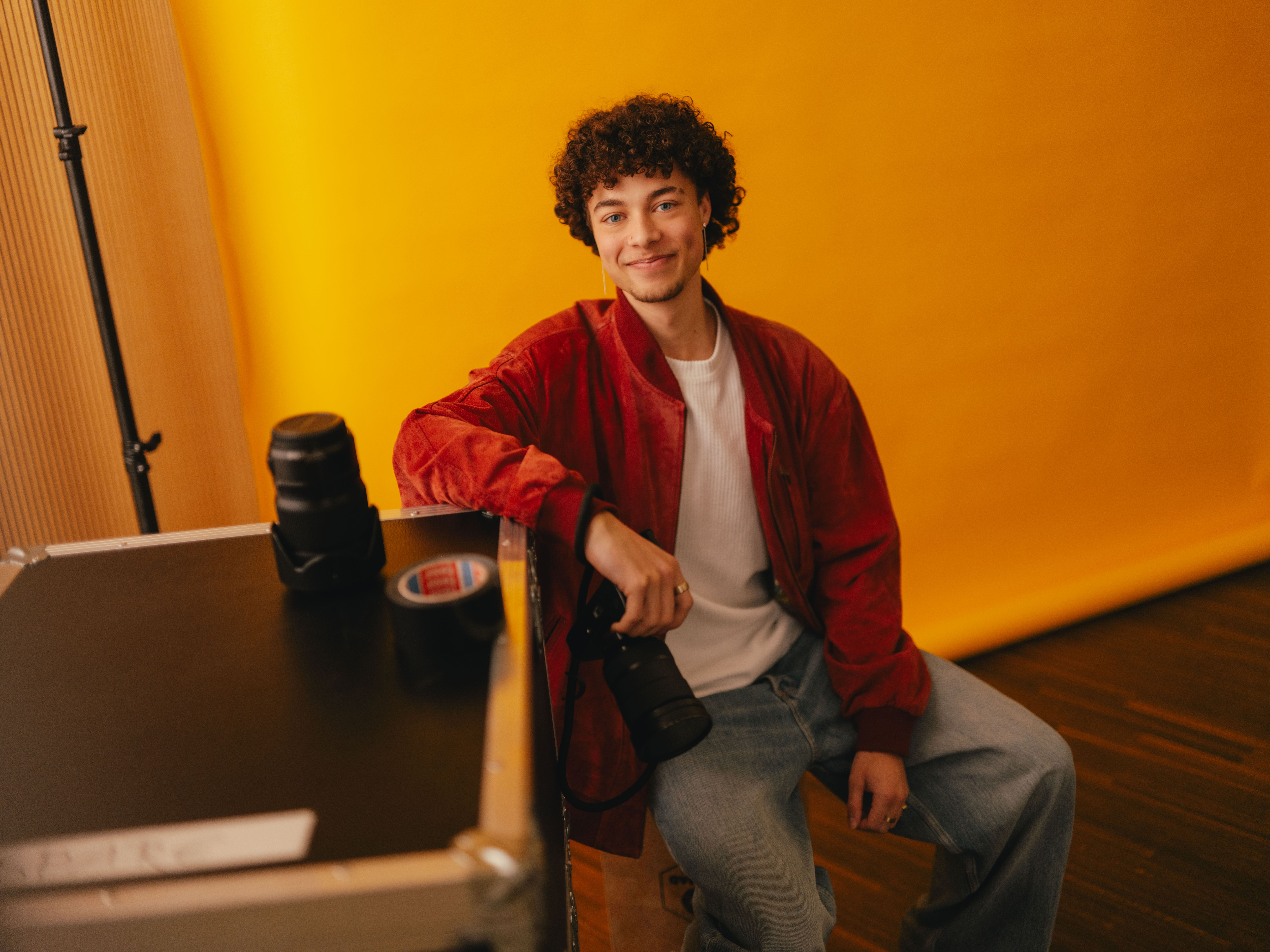 Una persona siede su una valigetta per il trasporto davanti a uno sfondo giallo brillante. Sulla valigetta ci sono obiettivi per la fotocamera e un rotolo di nastro adesivo. La persona indossa una giacca rossa, una t-shirt bianca e un paio di jeans larghi e tiene una macchina fotografica in mano. La sala ha l’aspetto di uno studio fotografico con attrezzatura professionale.