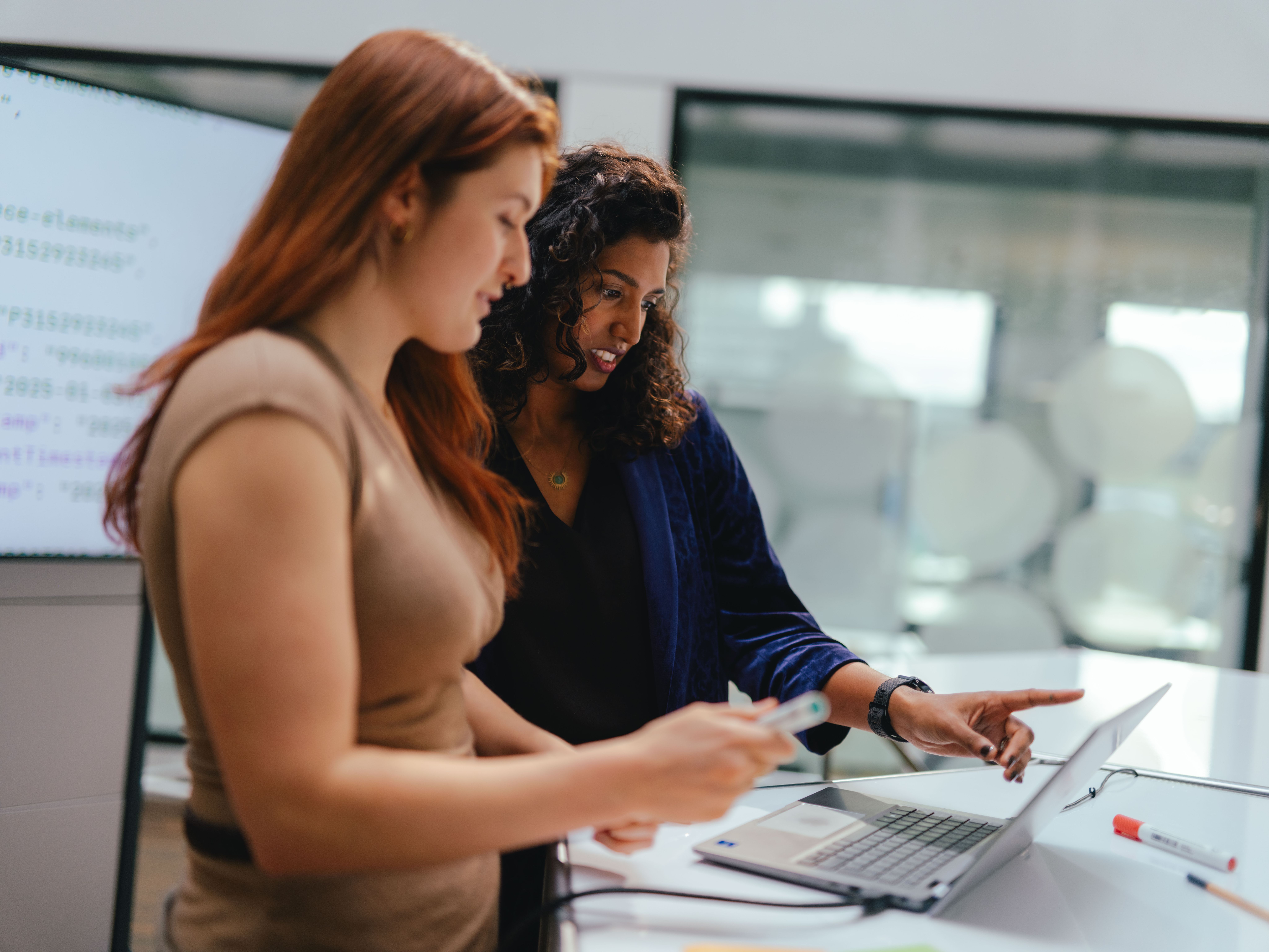 Deux personnes se tiennent à une table dans un espace de bureau moderne et travaillent ensemble sur un ordinateur portable ouvert. L’une tient un stylo ou un objet similaire, tandis que l’autre pointe vers l’écran. Sur la table se trouvent d’autres accessoires de travail, notamment un câble et un marqueur. À l’arrière‑plan, on voit de grandes fenêtres et des parois vitrées.