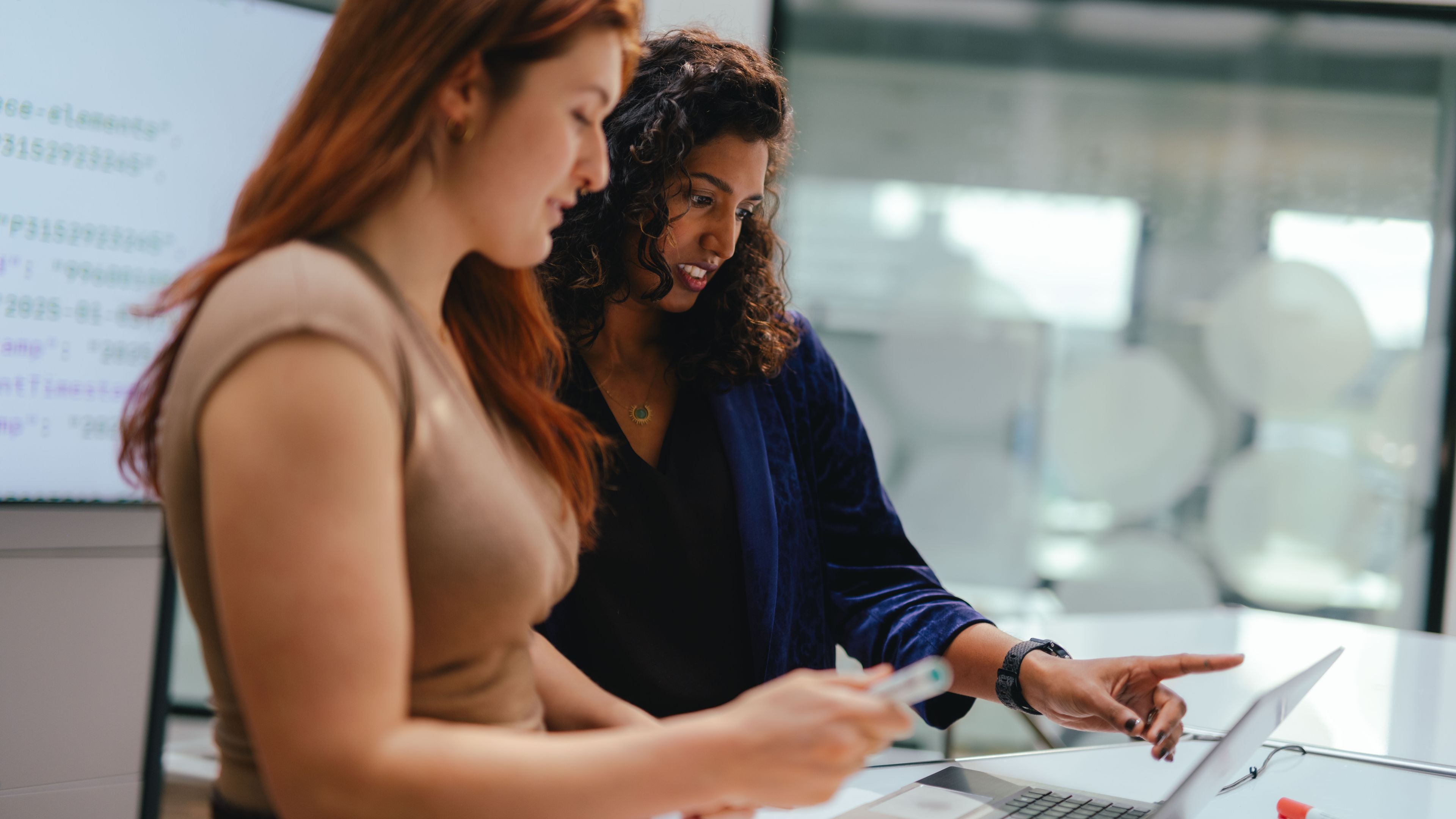 Two people are standing at a table in a modern office setting, working together on an open laptop. One person is holding a pen or similar object, while the other is pointing toward the screen. Additional work materials, including a cable and a marker, are placed on the table. Large windows and glass partitions are visible in the background.
