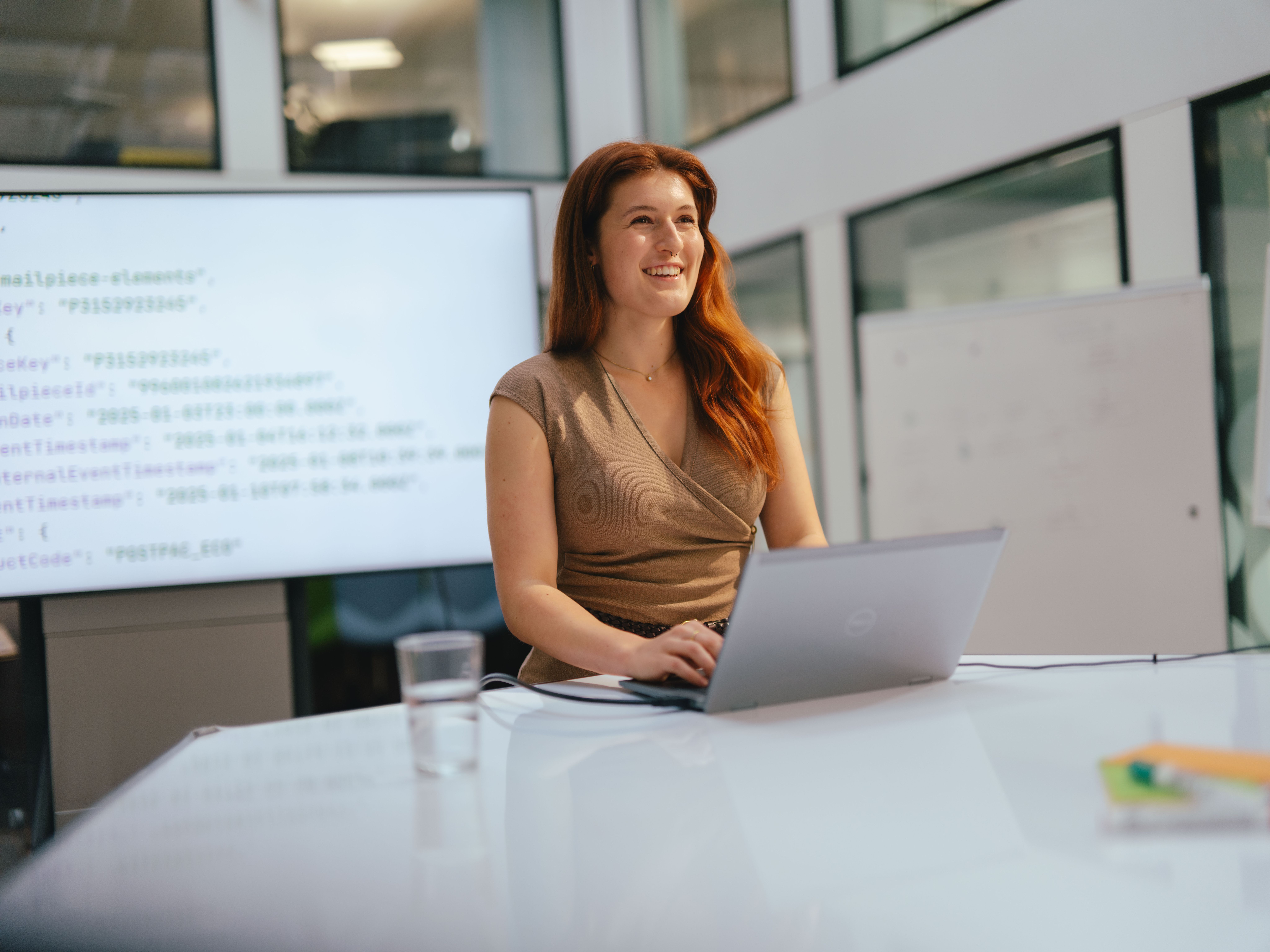 Une personne assise à une table avec un ordinateur portable ouvert tape sur le clavier. Un grand écran avec un document ouvert ainsi qu’un tableau d’affichage blanc se trouvent en arrière-plan. Un verre d’eau et d’autres matériaux de travail se trouvent également sur la table. L’espace est moderne et lumineux, avec de grandes fenêtres et des lignes claires.