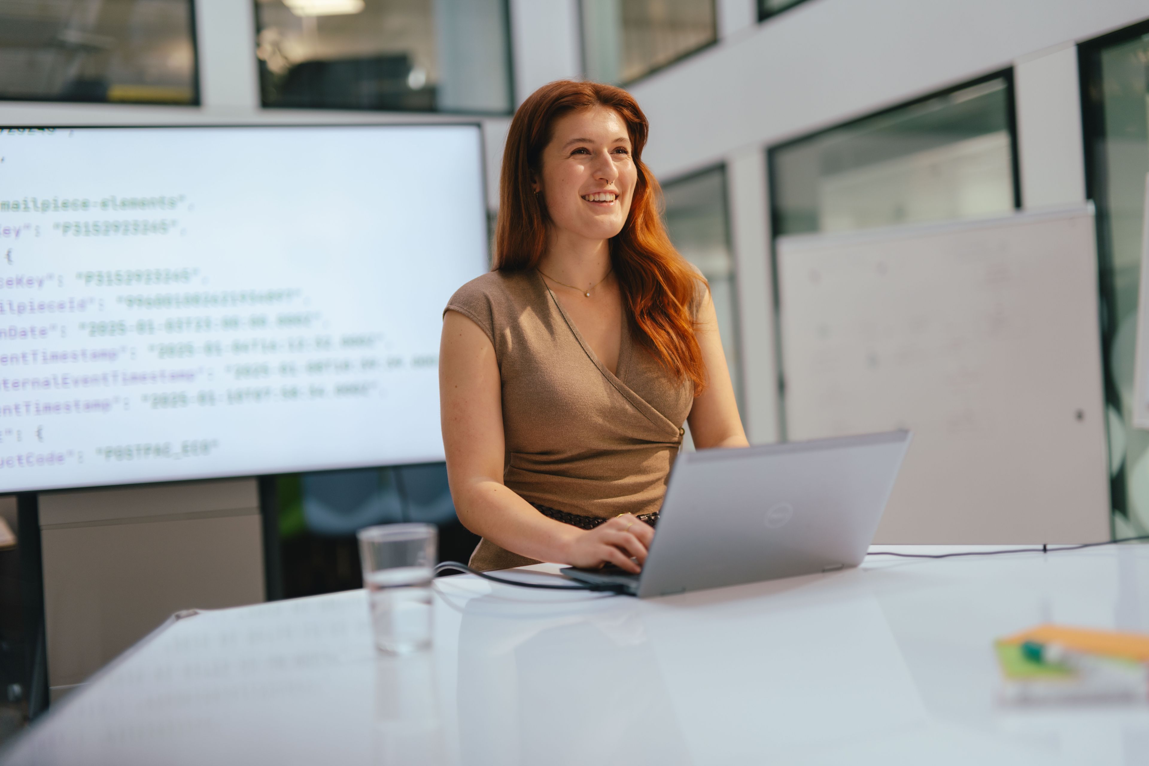 Une personne assise à une table avec un ordinateur portable ouvert tape sur le clavier. Un grand écran avec un document ouvert ainsi qu’un tableau d’affichage blanc se trouvent en arrière-plan. Un verre d’eau et d’autres matériaux de travail se trouvent également sur la table. L’espace est moderne et lumineux, avec de grandes fenêtres et des lignes claires.