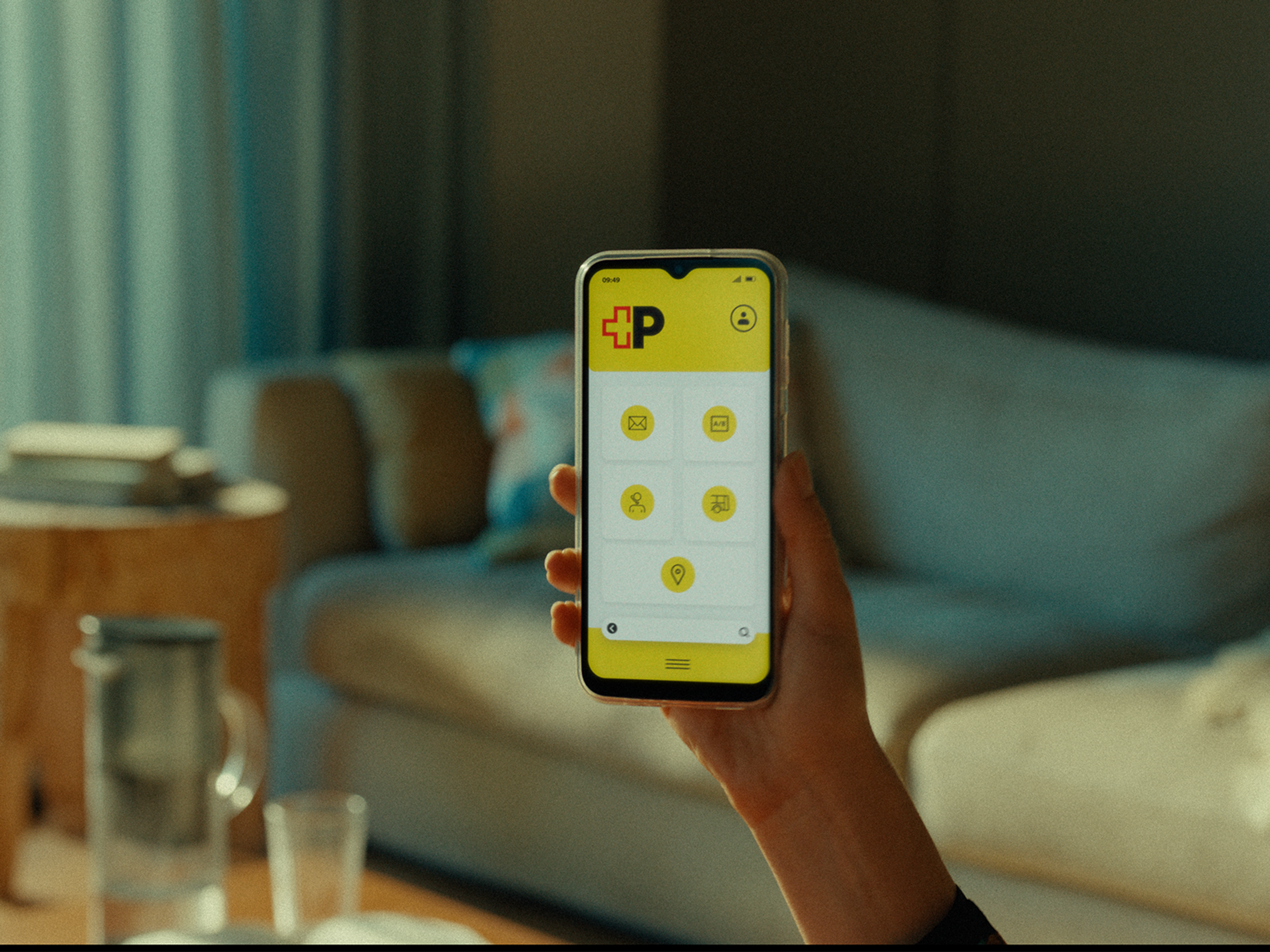 A hand holds a smartphone in front of a sofa in a living room. The phone screen displays a yellow interface with the red‑and‑black postal logo and several circular icons. In the background, a sofa, a table, and household items are visible.