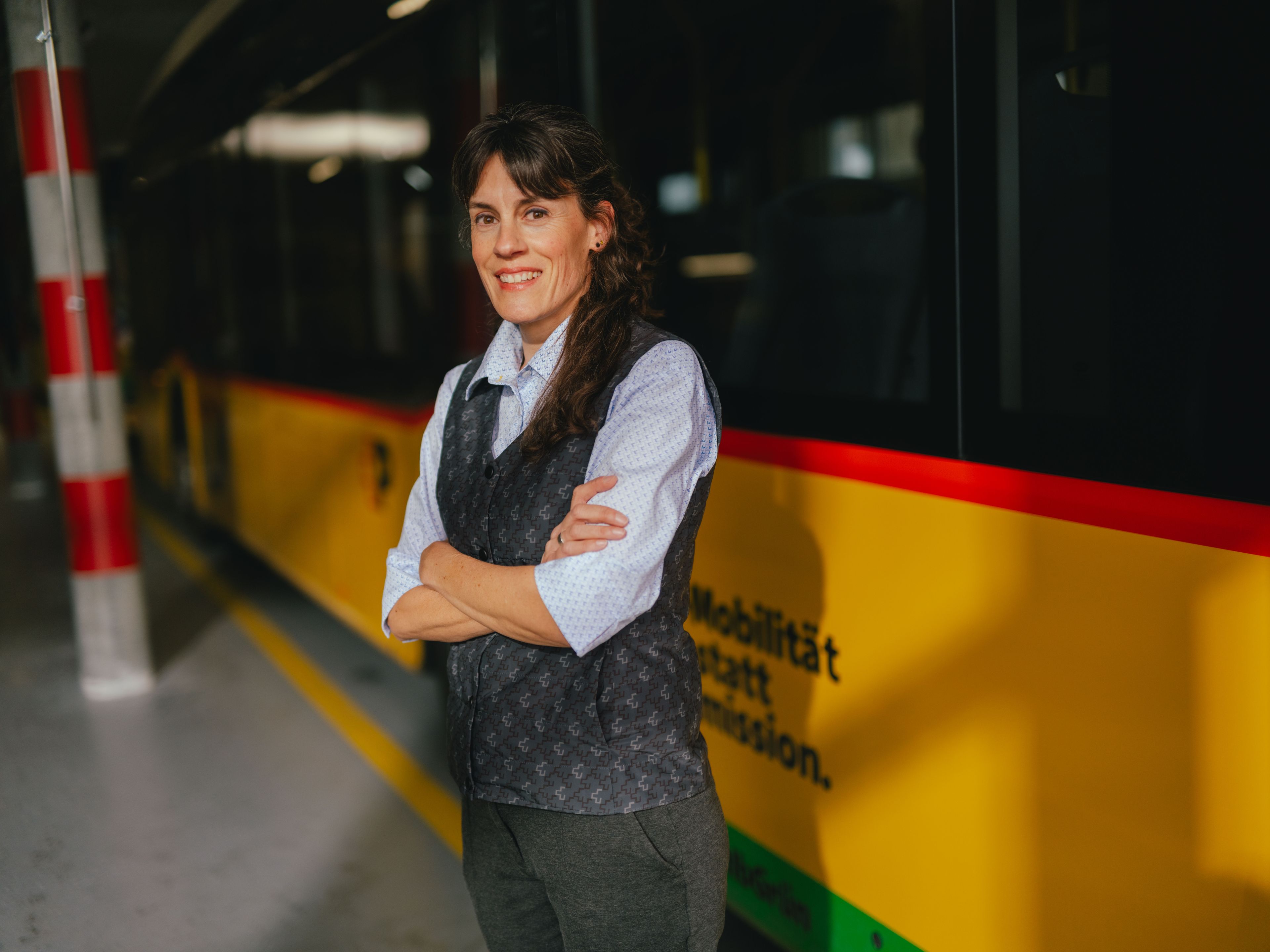 A person stands with arms crossed in a hall next to a yellow and black vehicle. The person is wearing a light shirt and a dark vest. The setting appears to be a depot or storage area, featuring striped pillars and a reflective floor.