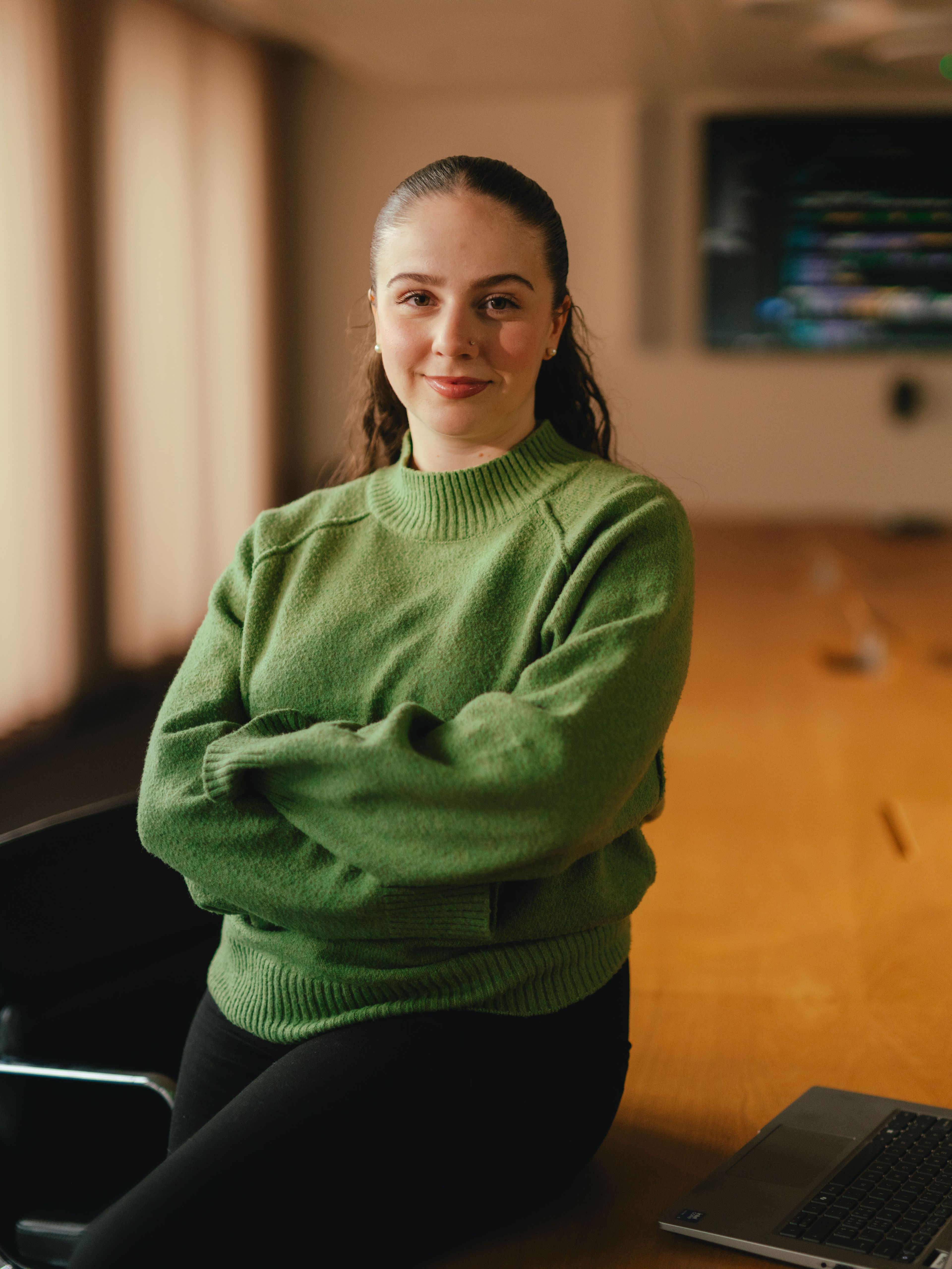 A person is standing in a conference room next to a large wooden table, arms loosely folded in front of their body. The person is wearing a green pullover. In the background, there is a large screen on the wall, several chairs and large windows with closed curtains. There is an open laptop on the table.