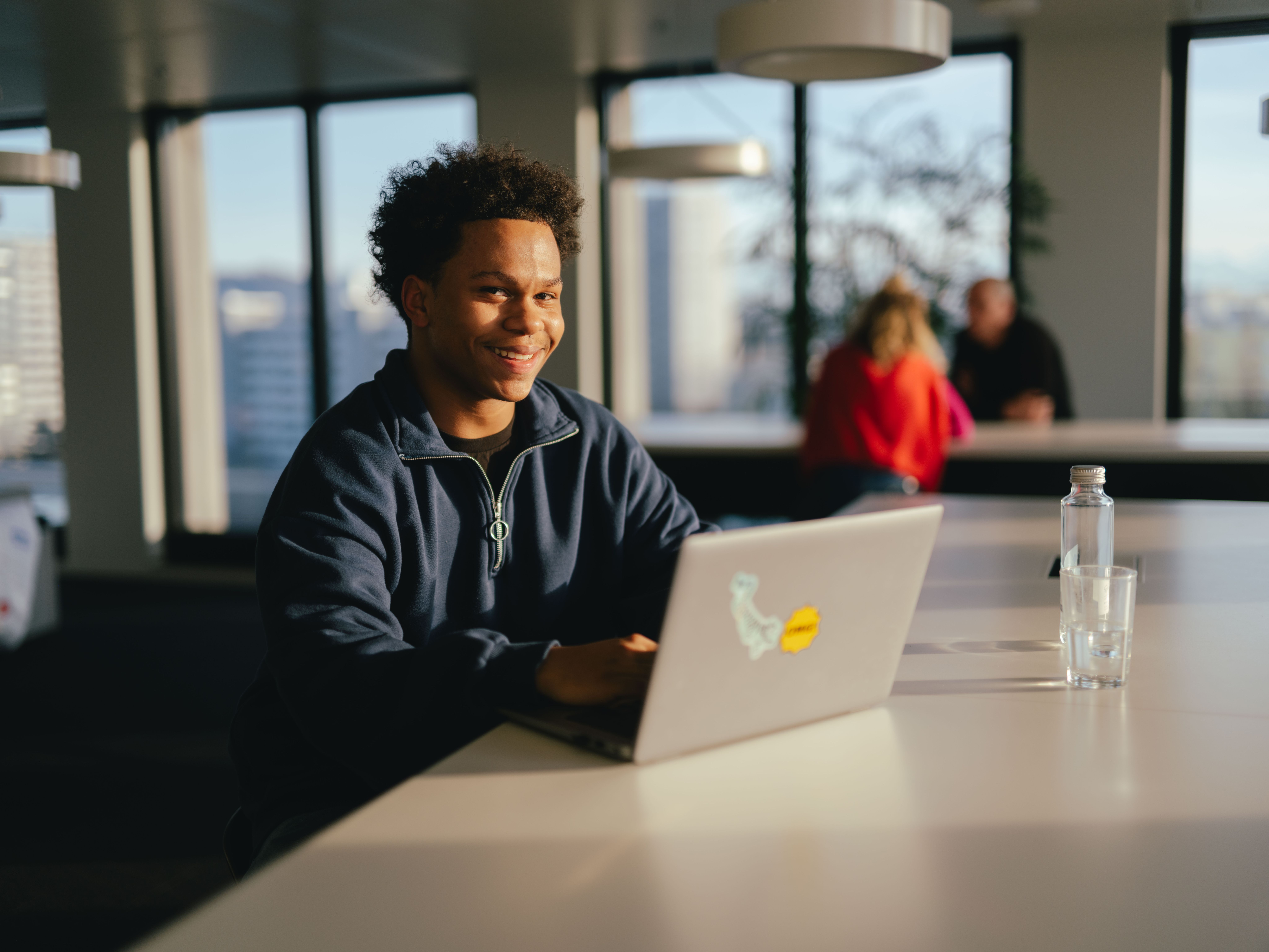 Eine Person sitzt an einem langen weissen Tisch und arbeitet an einem aufgeklappten Laptop, auf dessen Rückseite ein farbiges Aufklebermotiv zu sehen ist. Auf dem Tisch steht ausserdem eine durchsichtige Wasserflasche und ein leeres Glas. Der Raum ist hell, mit grossen Fenstern, durch die Tageslicht fällt. Im Hintergrund sind weitere Personen an einem Tisch sowie Büroelemente und Lampen sichtbar.