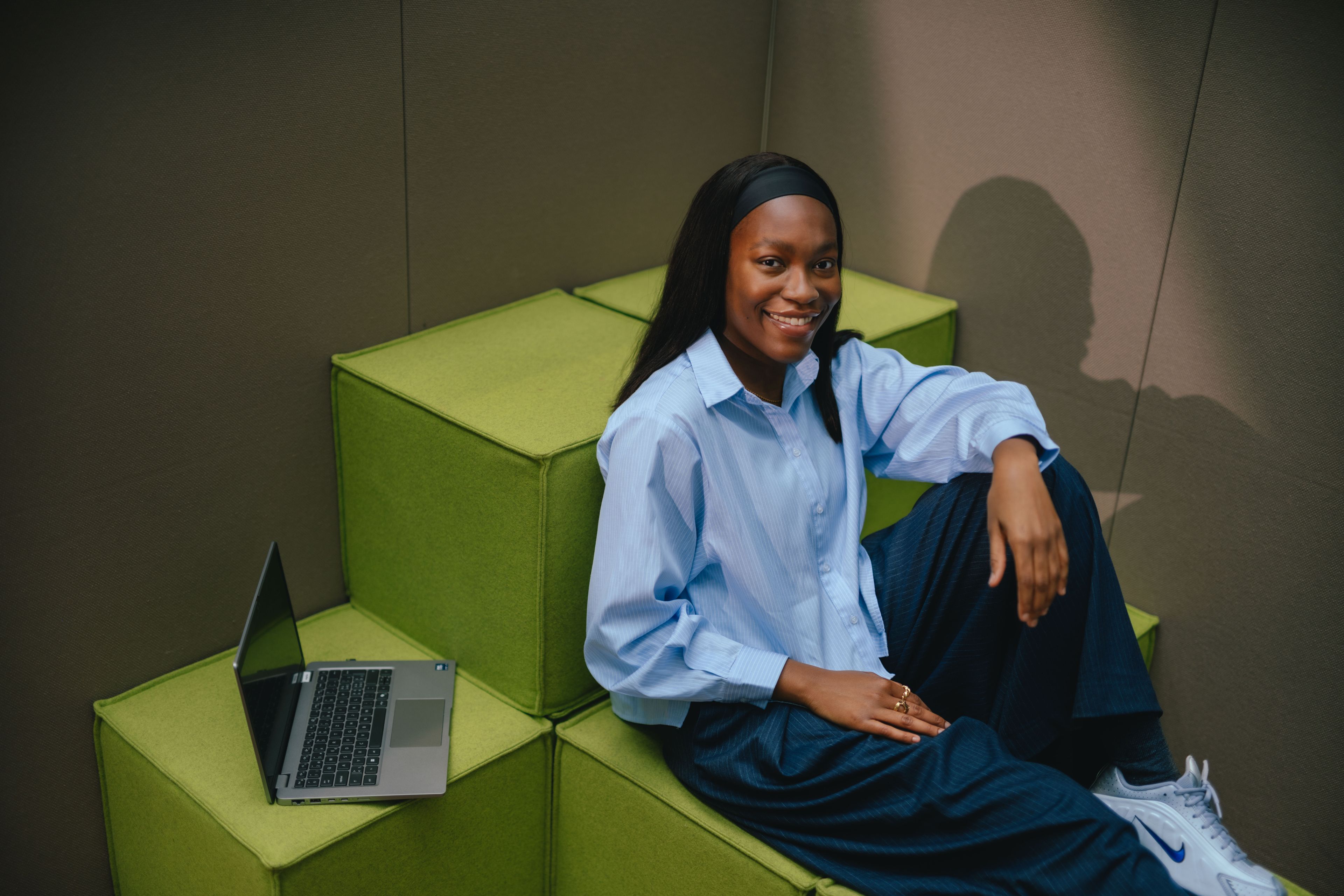 Una persona siede a un tavolo davanti a un laptop aperto. La persona si appoggia leggermente mentre tiene una mano sul laptop. Lo sfondo è composto da tende lunghe a terra e di colore beige.