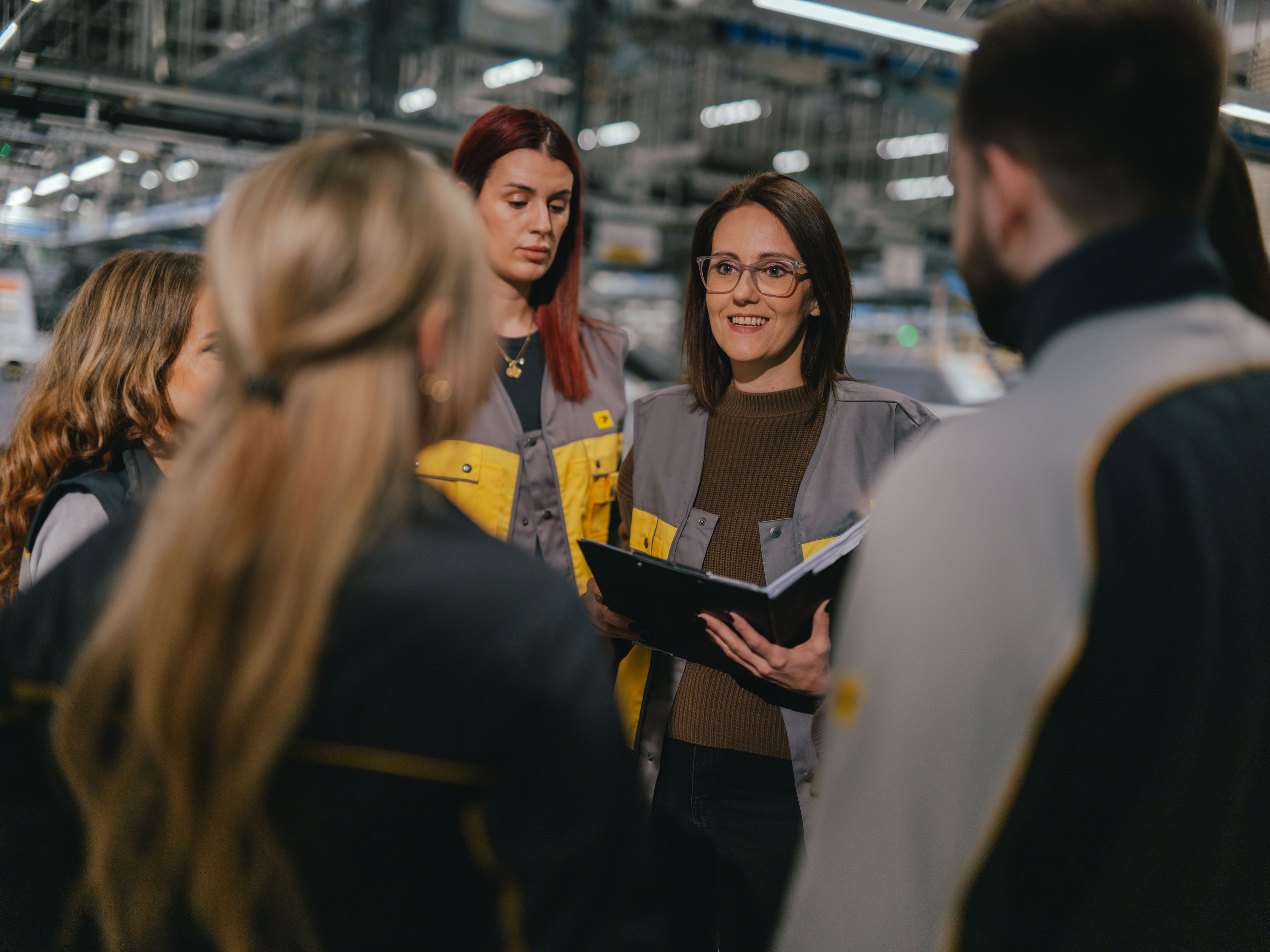 A group of people is gathered in a production or logistics hall. Several individuals are wearing grey and yellow work uniforms. One person is holding an open folder while the group stands in a circle. In the background, machines, shelving, and bright overhead lighting are visible.