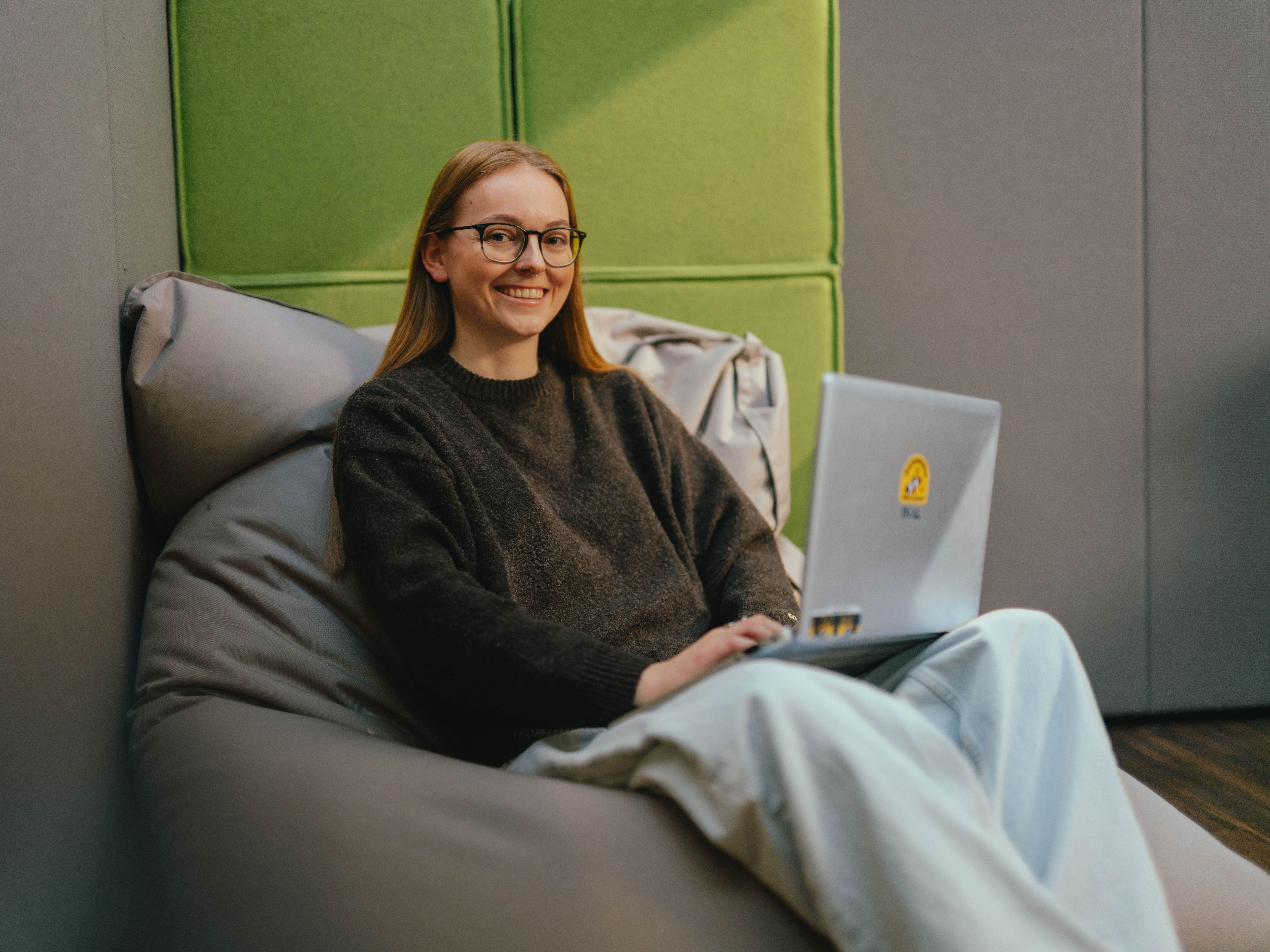 A person sits on a padded beanbag in a modern interior, working with a laptop folded open and lying on their lap. They are wearing a dark jumper and light, wide trousers. There are green padded wall panels behind the person. The space is designed to be quiet and feels like a retreat or work area.