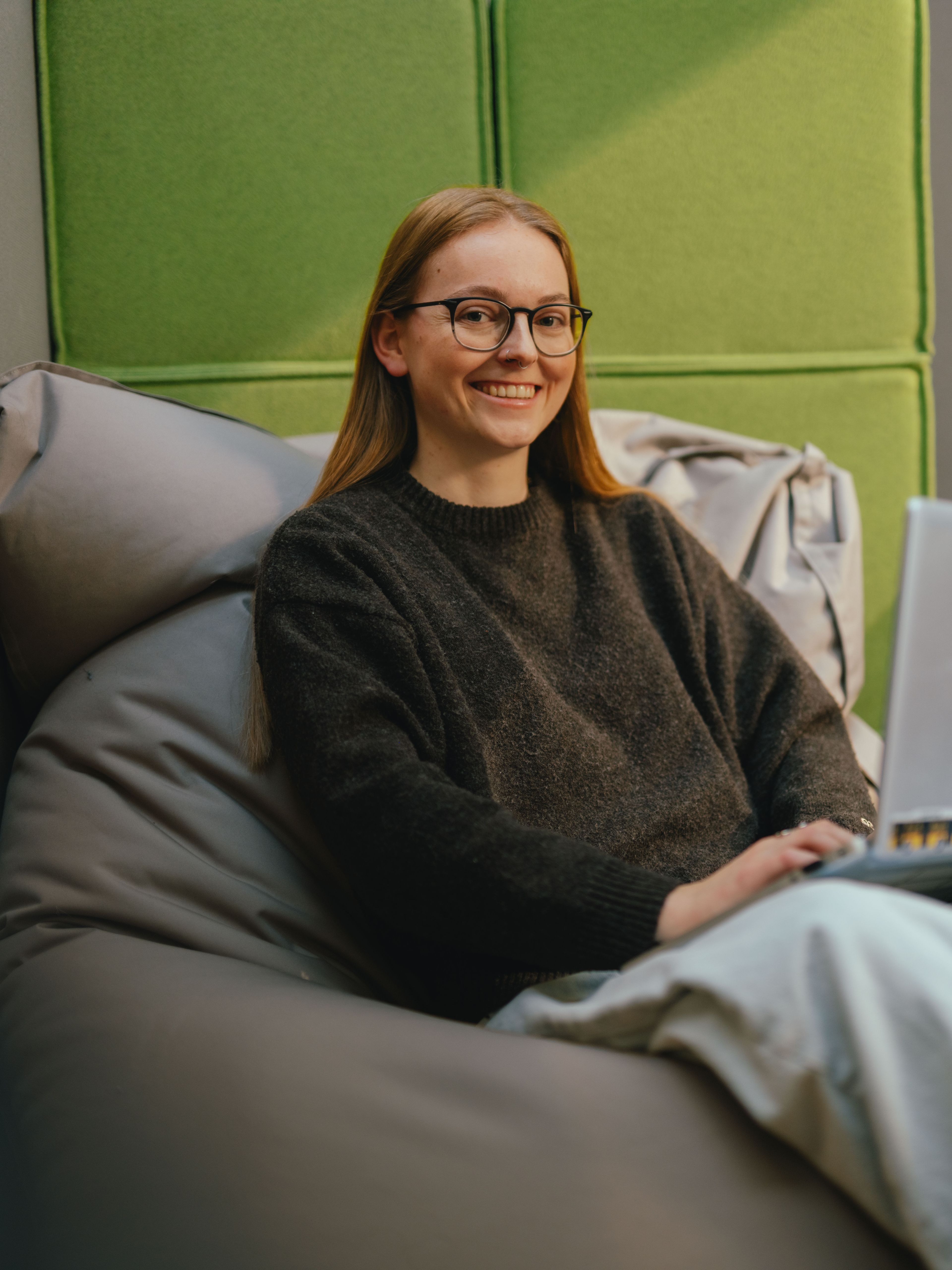 A person sits on a padded beanbag in a modern interior, working with a laptop folded open and lying on their lap. They are wearing a dark jumper and light, wide trousers. There are green padded wall panels behind the person. The space is designed to be quiet and feels like a retreat or work area.