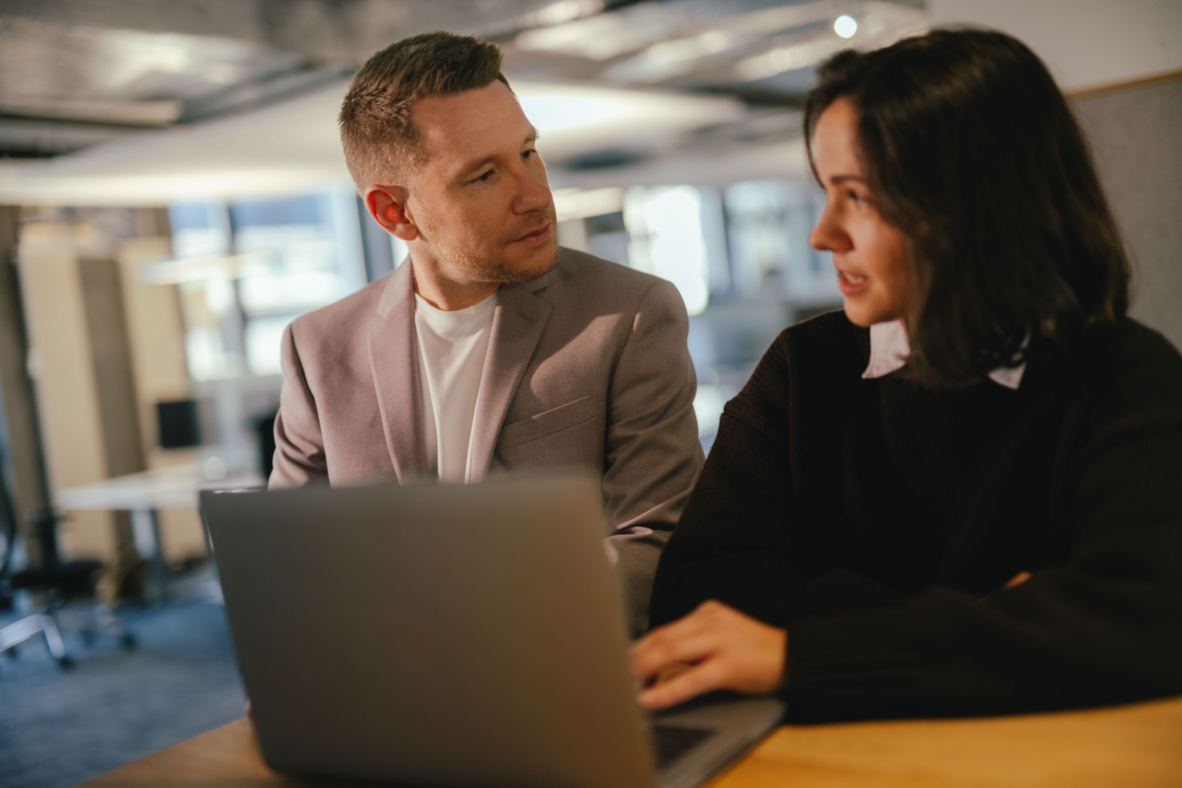 Zwei Personen sitzen an einem Tisch in einem modernen Büro und arbeiten gemeinsam an einem Laptop. Eine der Personen tippt auf der Tastatur, während die andere Person daneben sitzt und in Richtung des Bildschirms schaut. Im Hintergrund sind grosse Fenster und Büroeinrichtungen sichtbar.