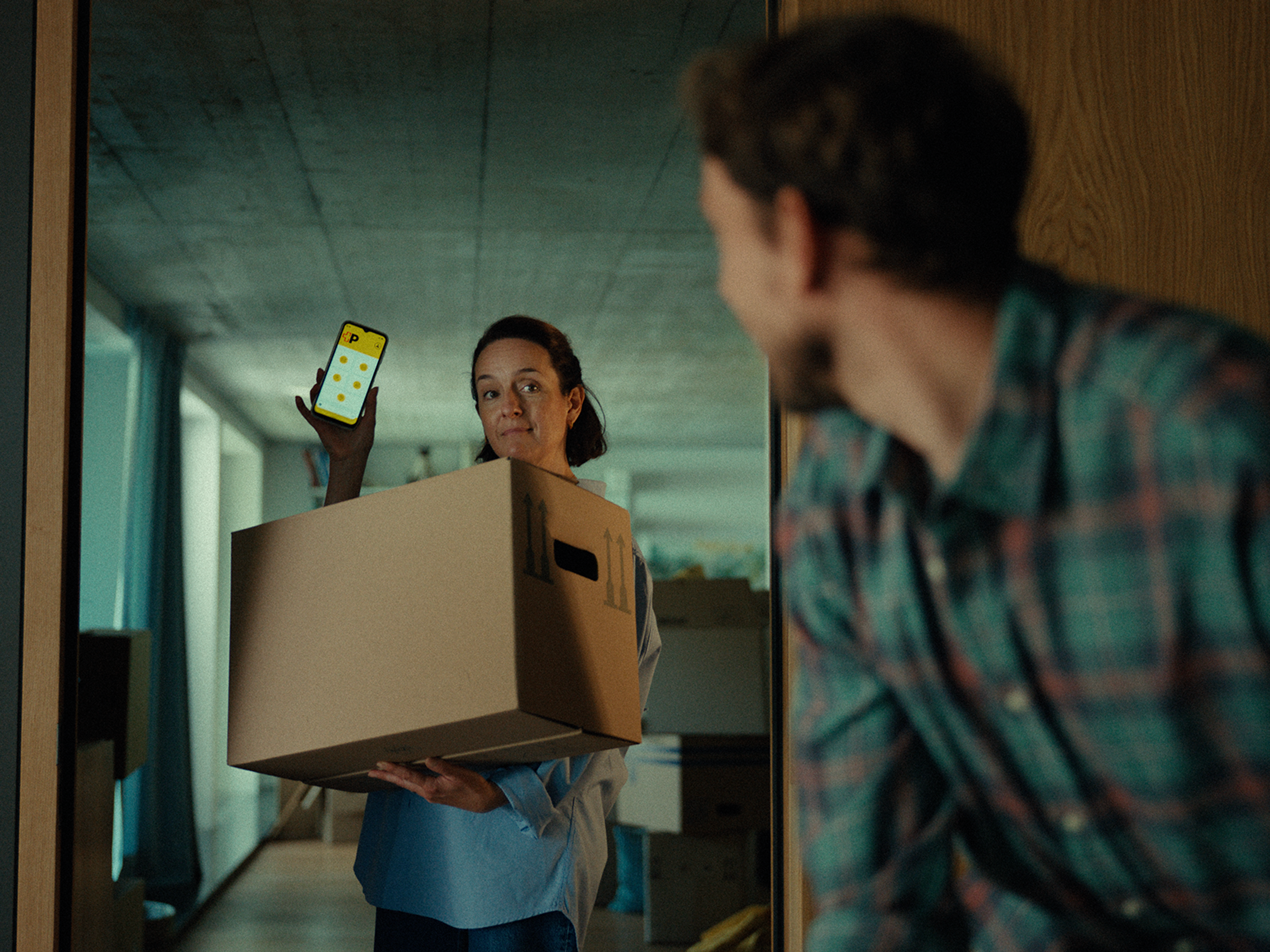 Inside a room, a person is carrying a large moving box in both arms while holding up a smartphone in the other hand. The phone screen displays a yellow interface with several icons. Another person is partially visible in the foreground from the side. The background includes furniture, boxes, and a hallway.