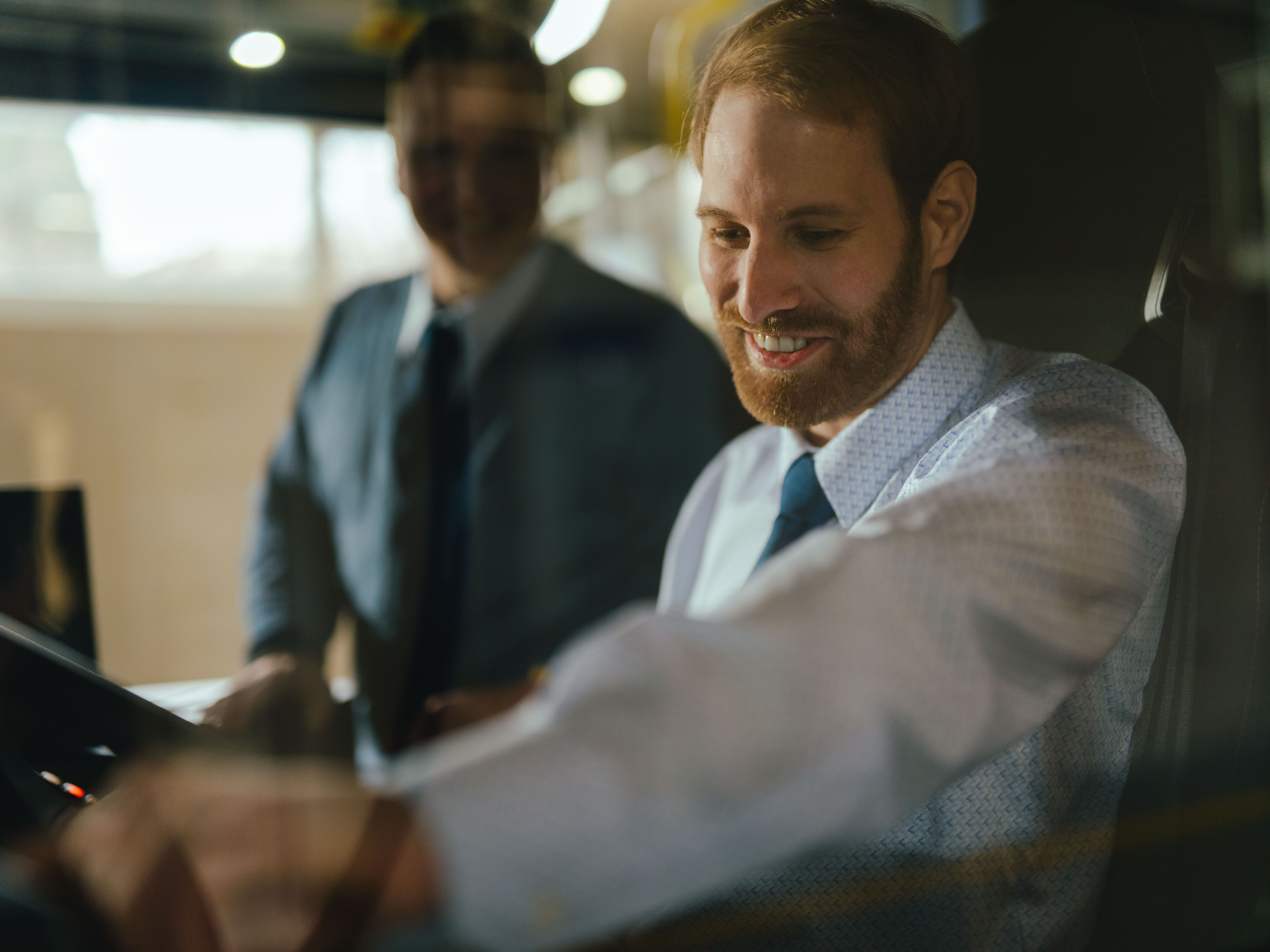 Two people are in the driver’s area of a vehicle. One person is seated at the wheel and reaching for a control, while the other stands beside them. The steering wheel, dashboard controls, and interior lighting are visible.